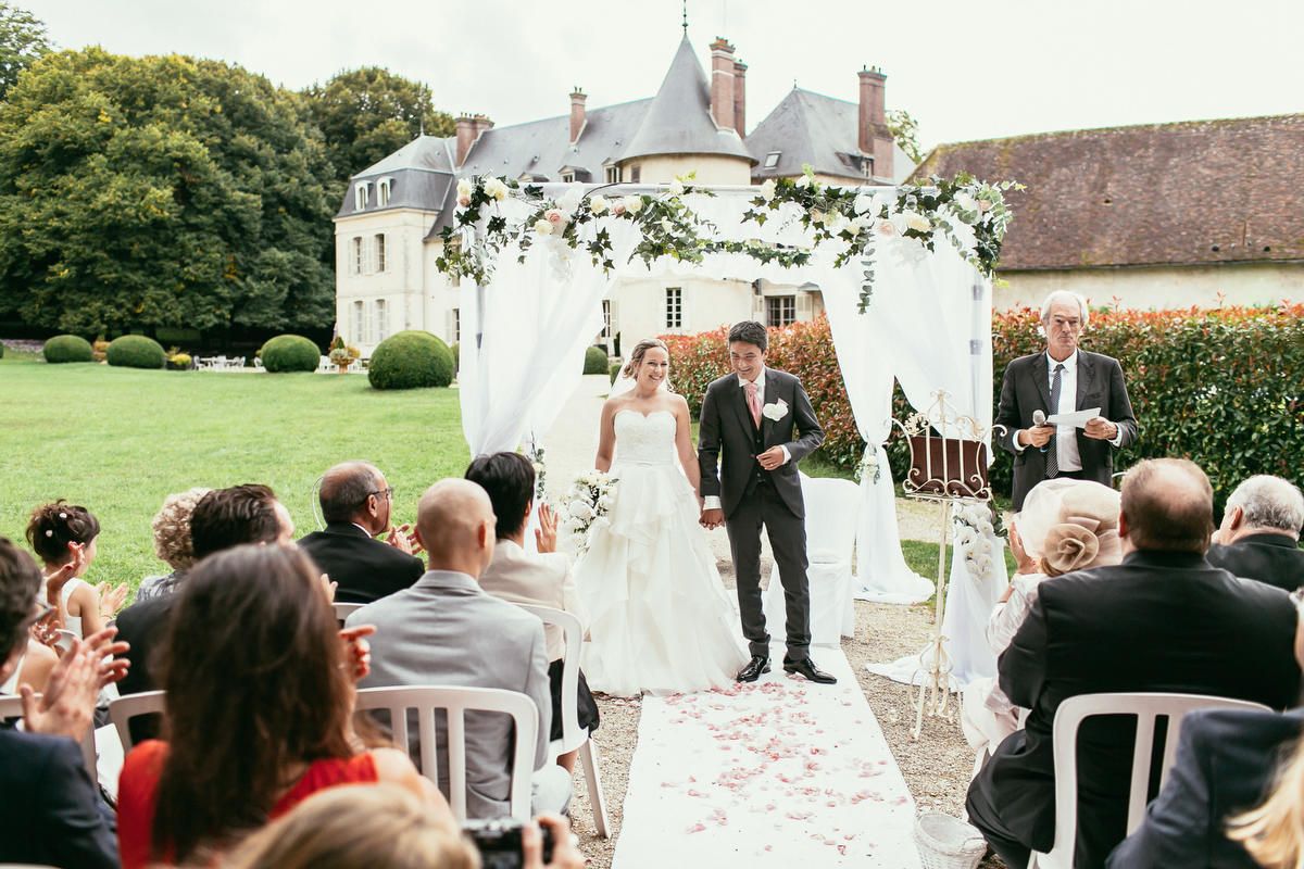 Bride and groom hold hands under a white floral arch during their outdoor chateau wedding ceremony near Paris as guests clap.