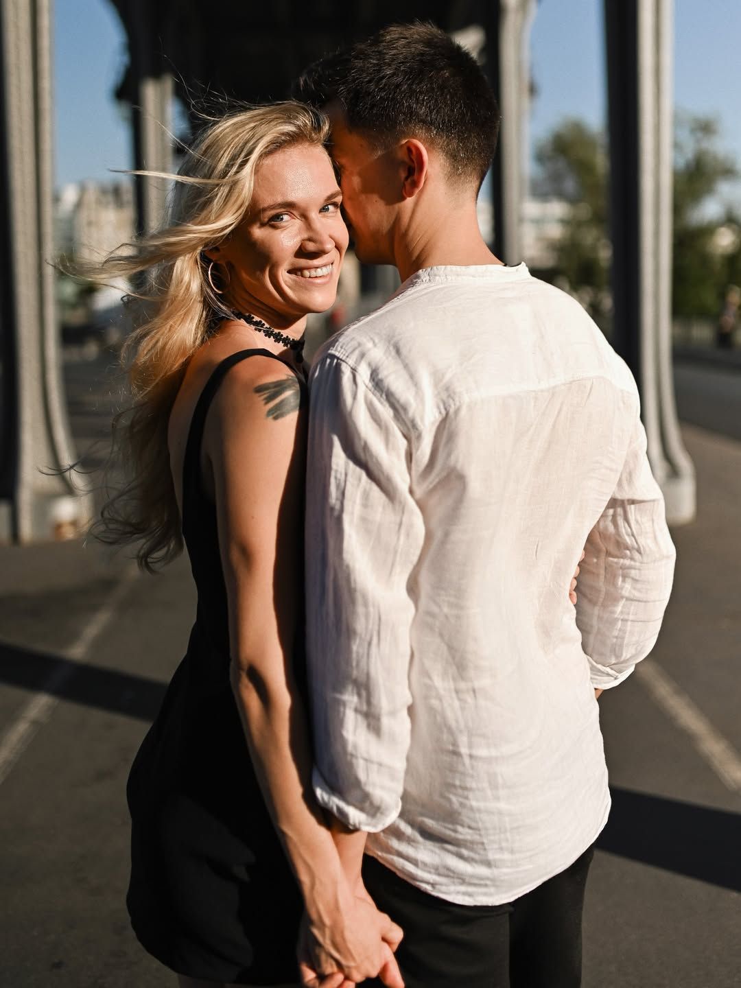 Smiling newly engaged woman hugging her partner under the metal pillars of Pont de Bir-Hakeim in warm golden hour sunlight.