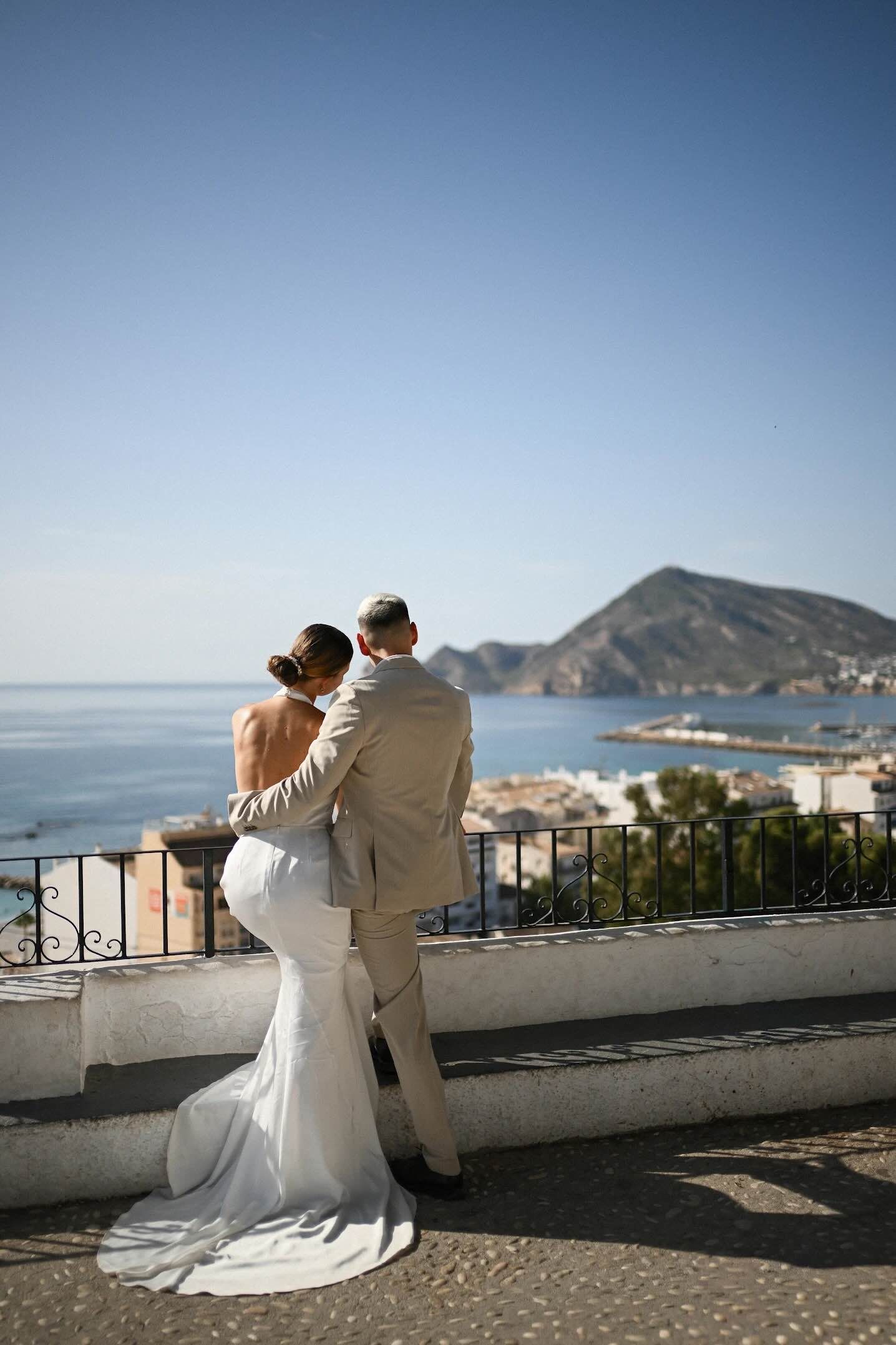 Newlyweds leaning on a white railing above Altea, looking out over the bay and the harbor on a clear afternoon.