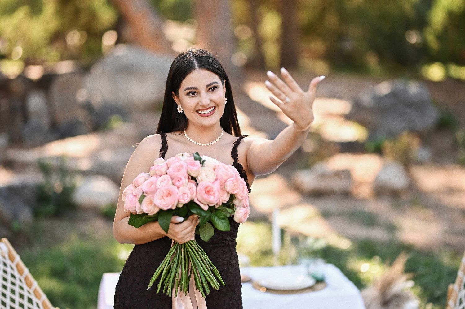 Happy bride-to-be holding a pink rose bouquet and showing off her new engagement ring to the camera in Viveros Park.