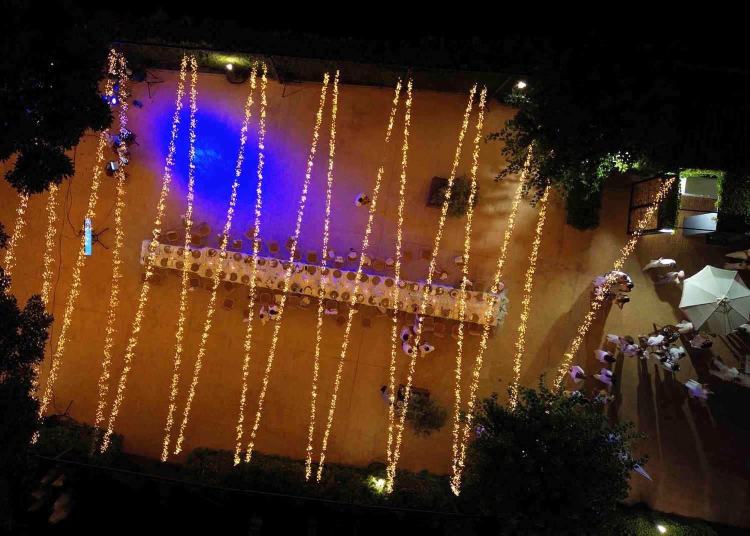 Top-down drone view of a long illuminated wedding banquet table and guests dancing outdoors at night in Spain.