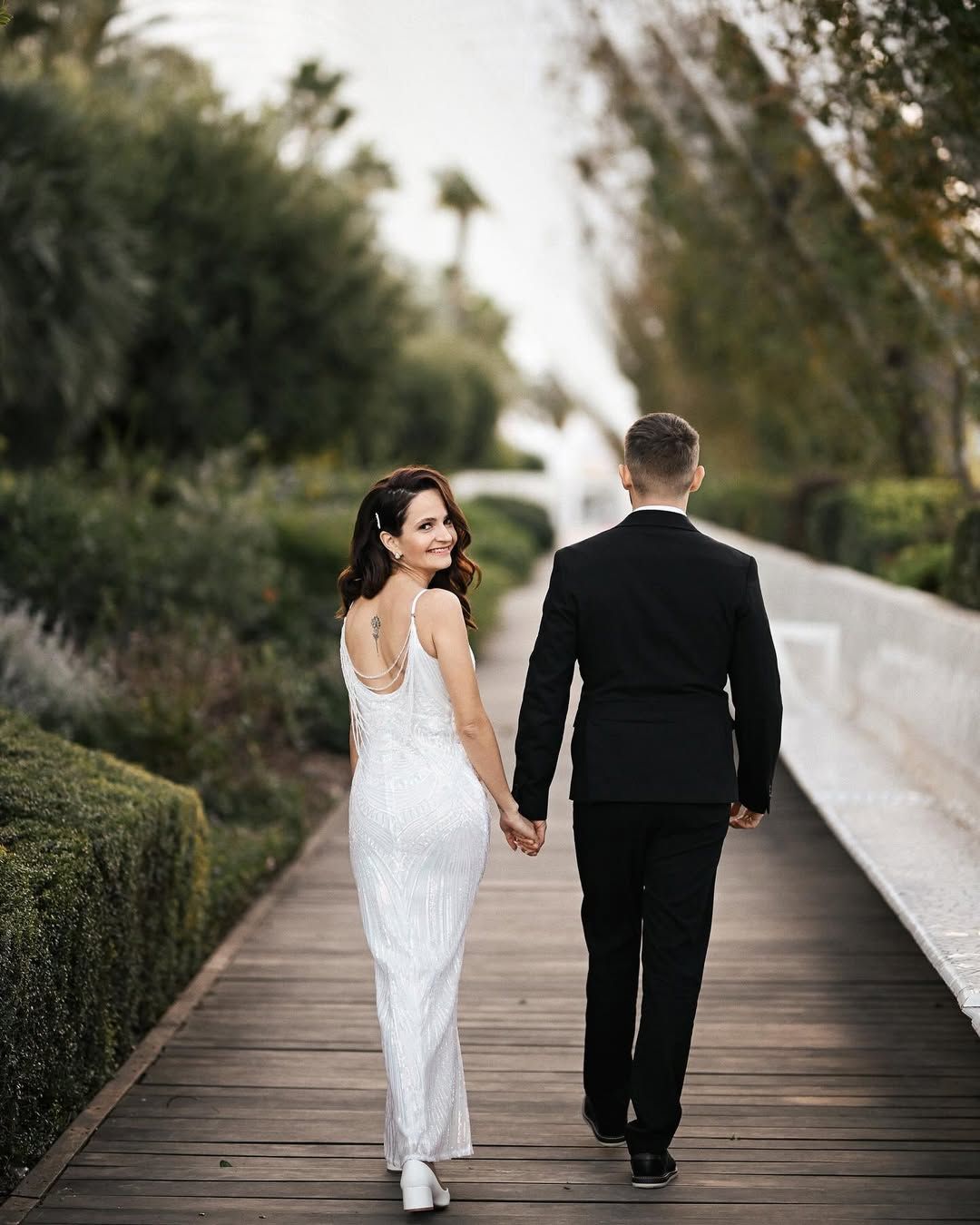 A bride in a white backless dress looks over her shoulder while walking with the groom through L'Umbracle gardens in Valencia.