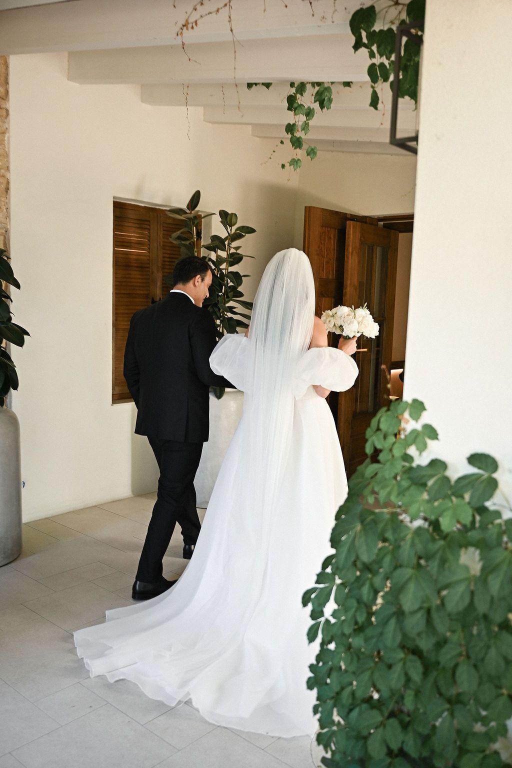 Full-length back view of the bride and groom walking to their ceremony, showcasing the flowing veil and long wedding dress train at the finc