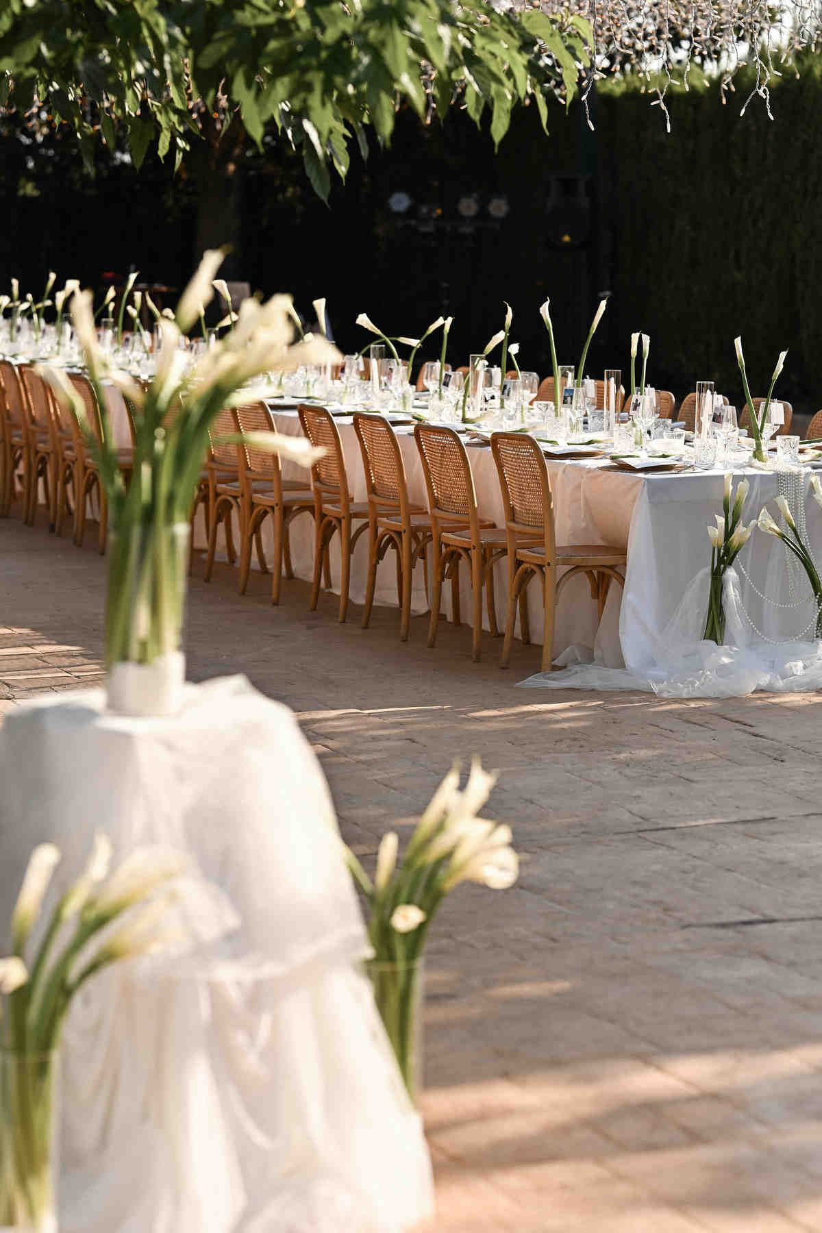Long outdoor wedding dinner table with wooden chairs and white calla lilies under a canopy of fairy lights.