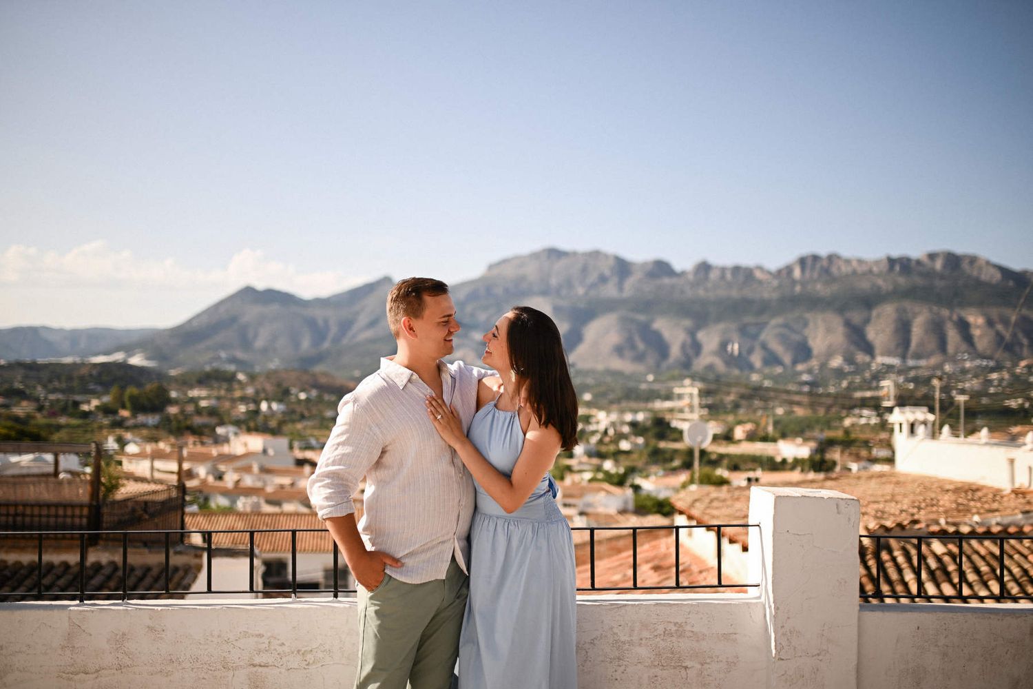An engaged couple stands on a white terrace looking at each other with the mountains and Altea Old Town in the background.