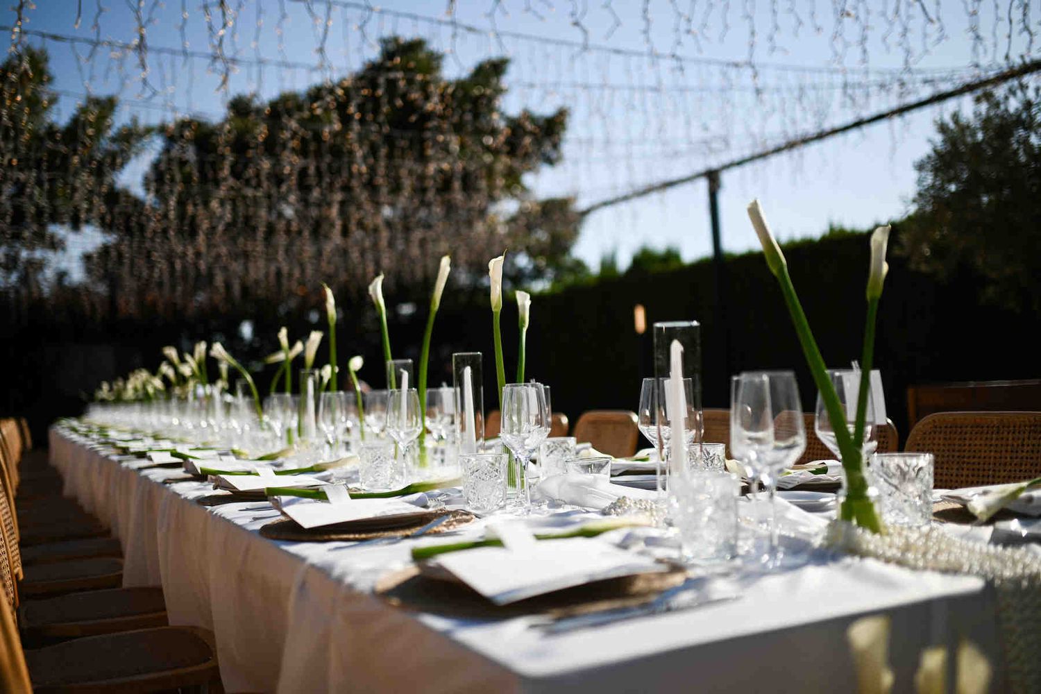 Close-up of crystal glasses, white candles, and elegant calla lily stems on a white tablecloth during golden hour.
