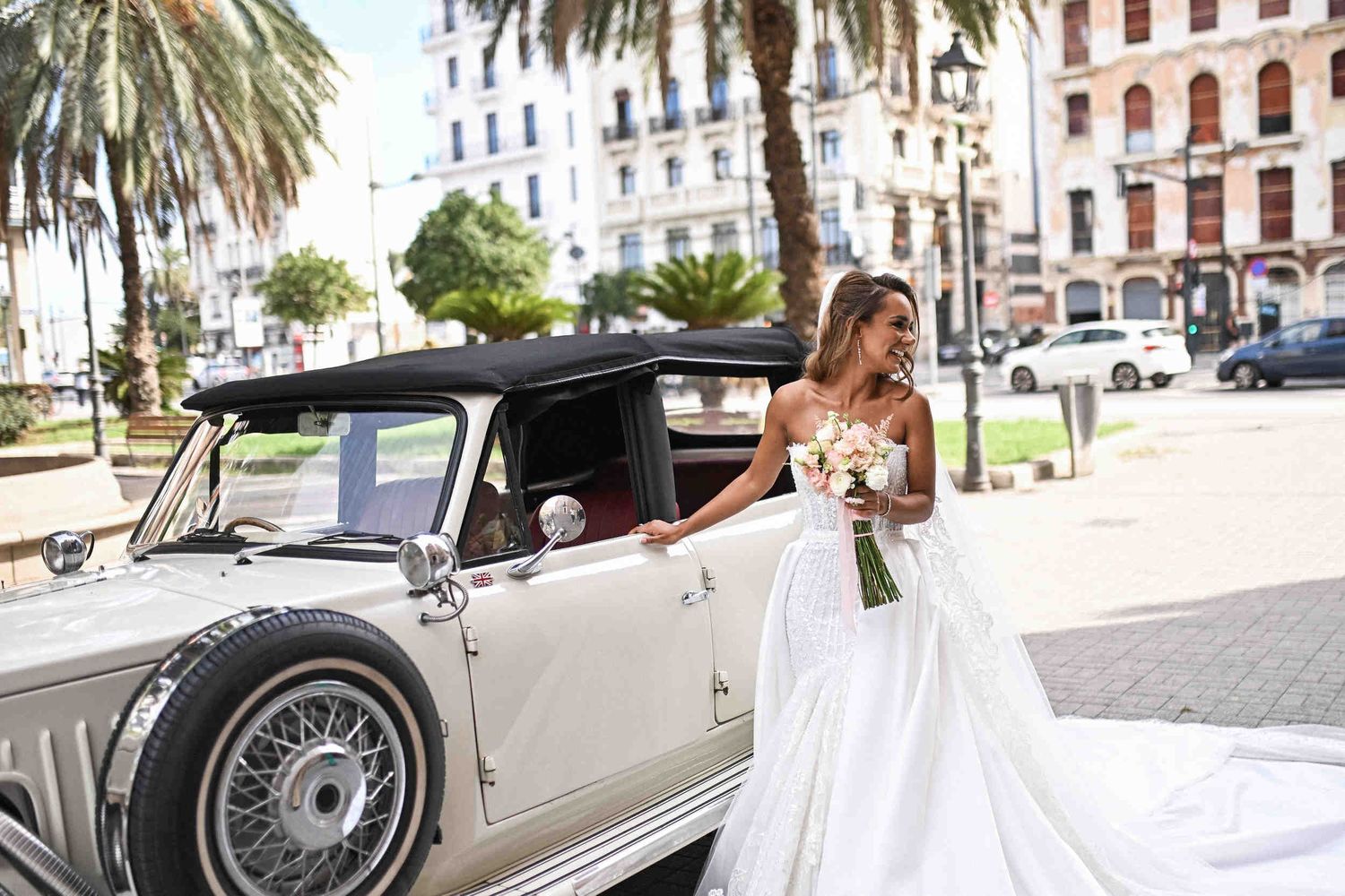 Smiling bride in a mermaid gown holding a bouquet next to a white vintage car outside Hotel Las Arenas.