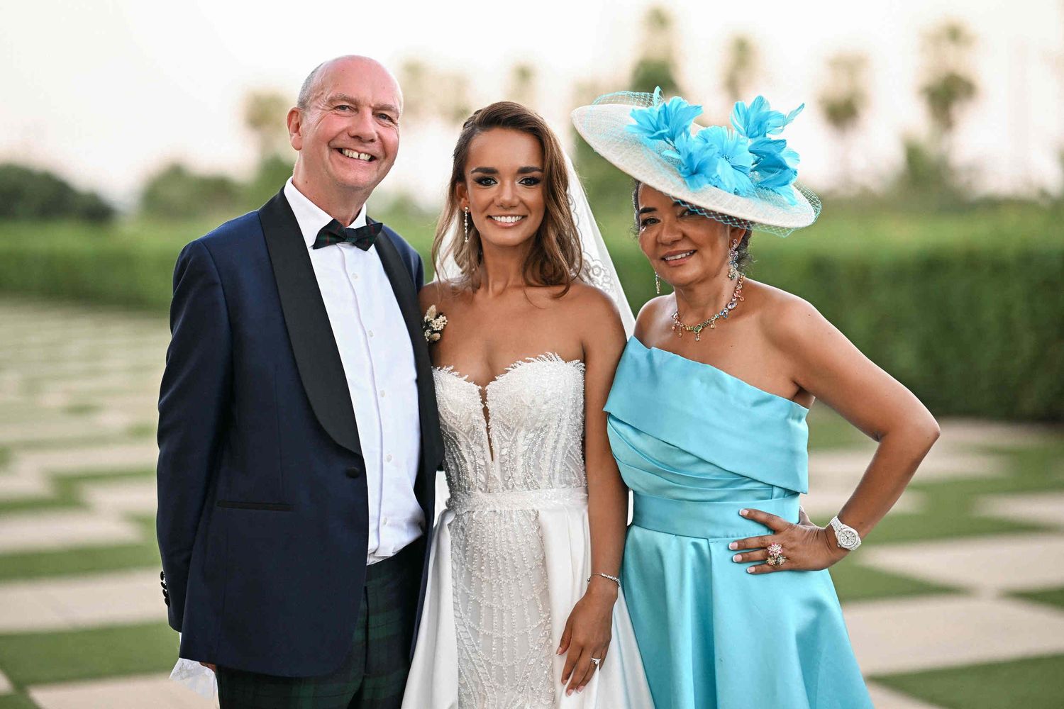 Bride smiling with her father in a tuxedo and mother in a blue hat during family photos at Hotel Las Arenas