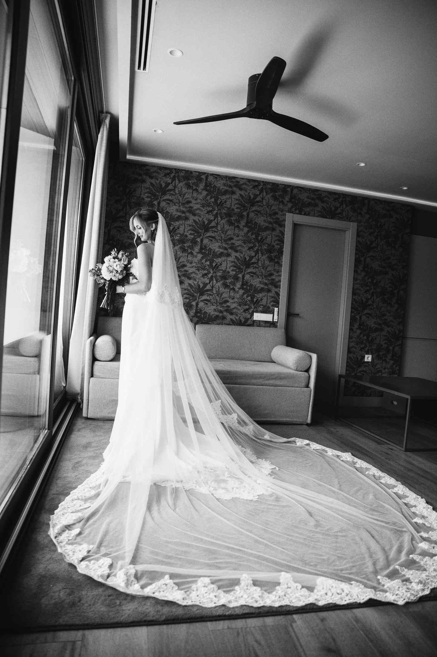 Black and white full-length portrait of a bride with a long flowing wedding veil inside a spacious hotel room Apartamentos Core Suites