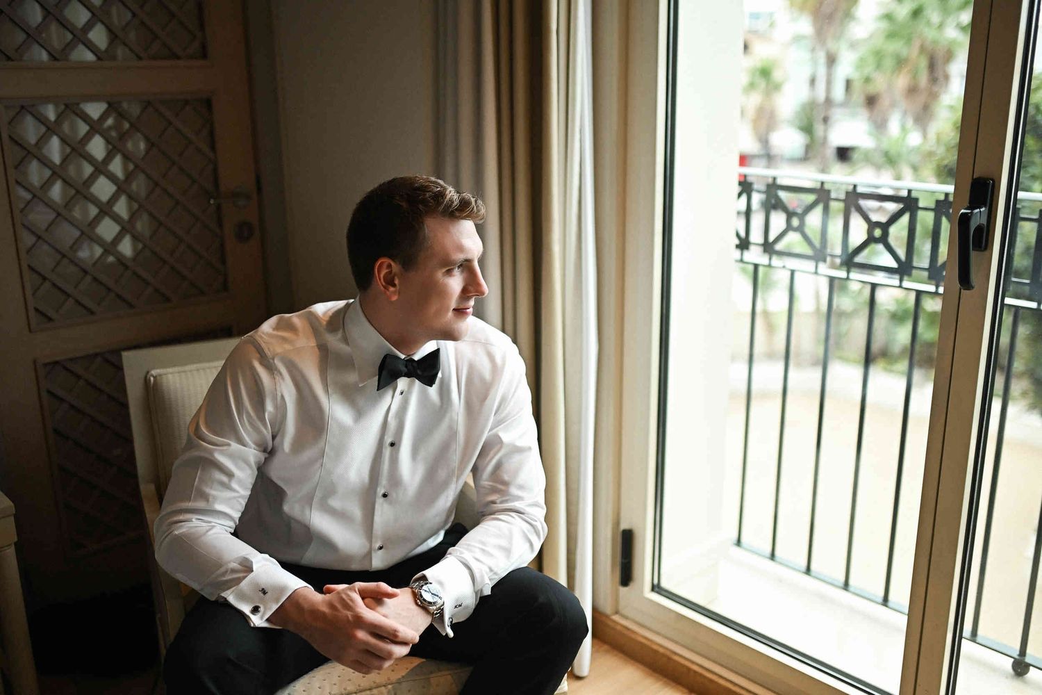 Groom wearing a white shirt and bowtie sitting thoughtfully by a large window in a luxury hotel suite.