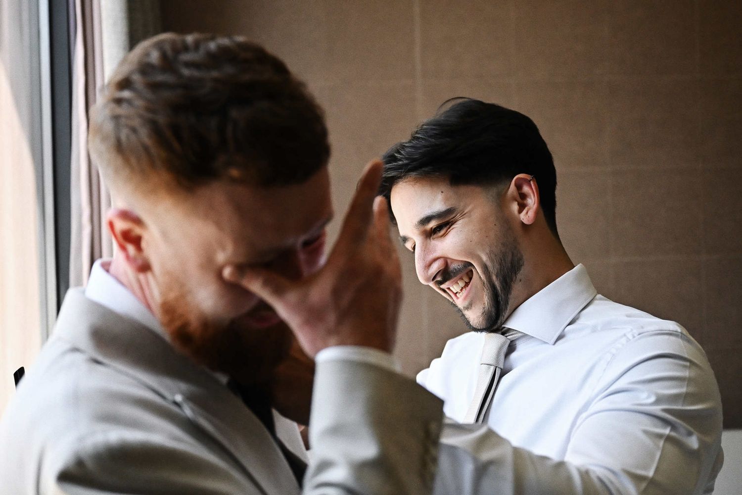 Candid moment of an emotional groom crying while his best man laughs and attaches the boutonniere in a hotel suite.