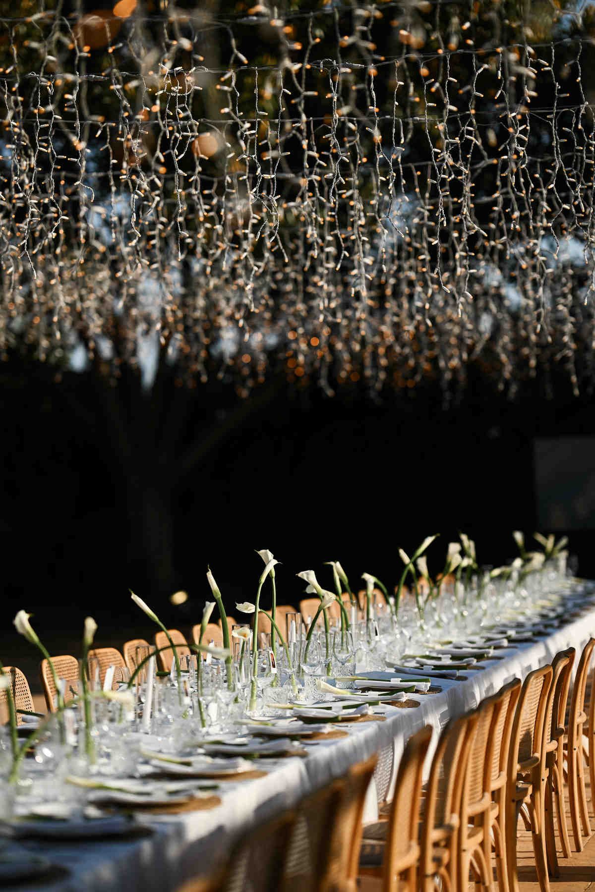 Warm golden hour light illuminating a long wedding banquet table with calla lilies and hanging fairy lights at Alquería Kukala.