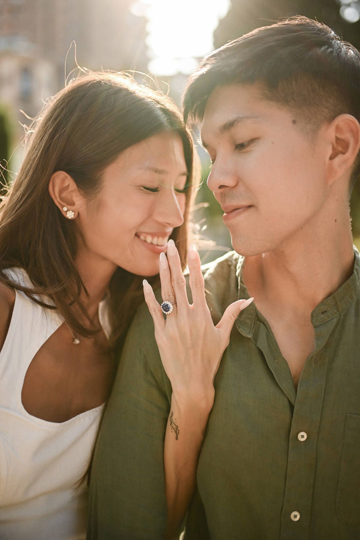 A woman displays her blue ring resting on her partner's chest in warm sun flare after a Jardines de Monforte proposal.