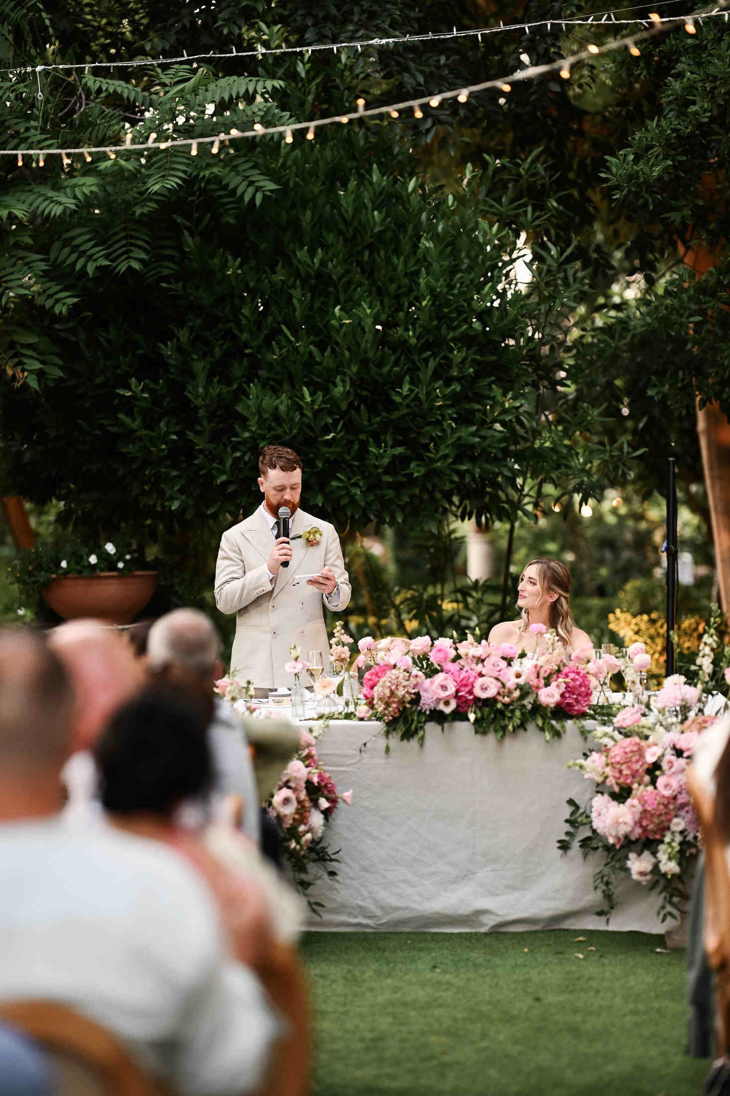 Groom speaking to guests showcasing the luxury wedding reception decor, head table podium, and beautiful outdoor lighting.