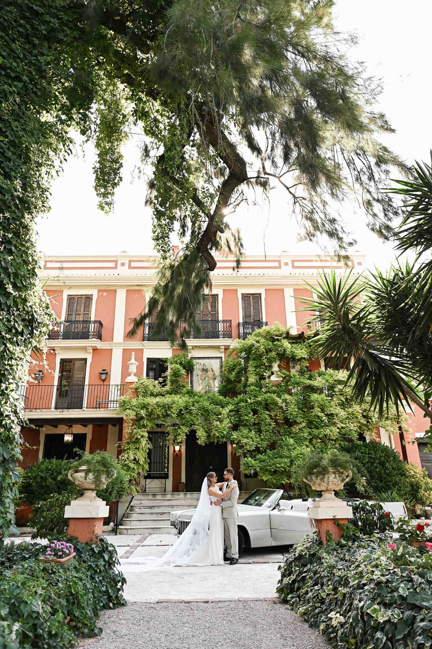 Wide shot of a bride and groom during a wedding photoshoot with a vintage retro car at the scenic Villa Dela estate.