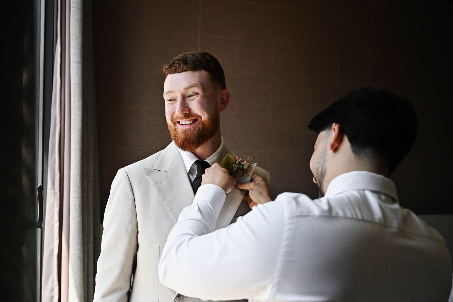 Best man helping the groom by attaching a stylish floral boutonniere to his suit jacket during preparations.