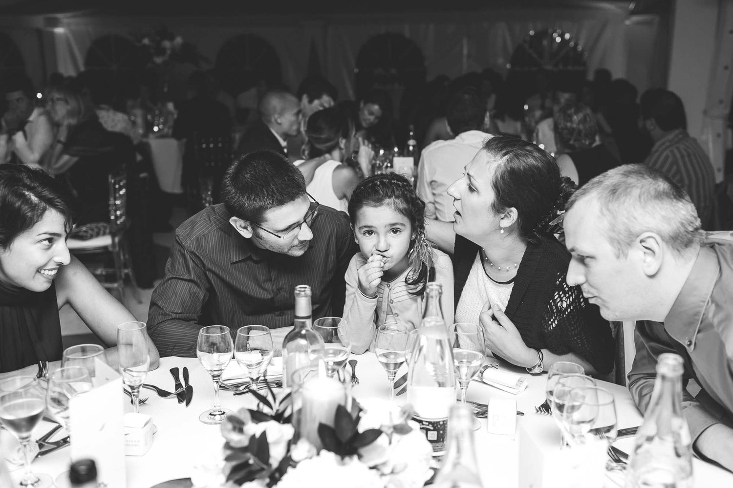 B&W documentary shot of a little girl looking at the photographer while guests socialize during a European wedding reception