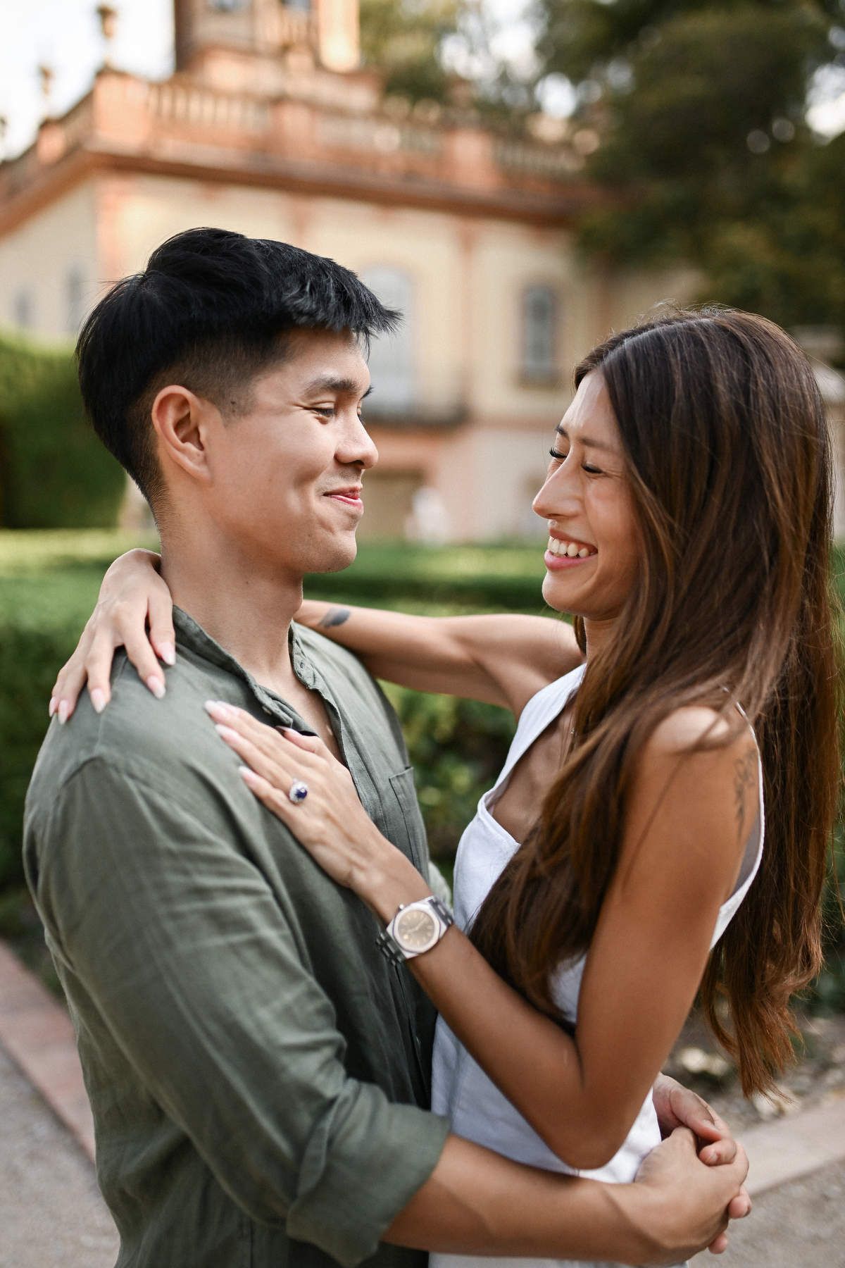 Happy engaged couple laughing and looking into each other's eyes, with classical Spanish architecture in the background.