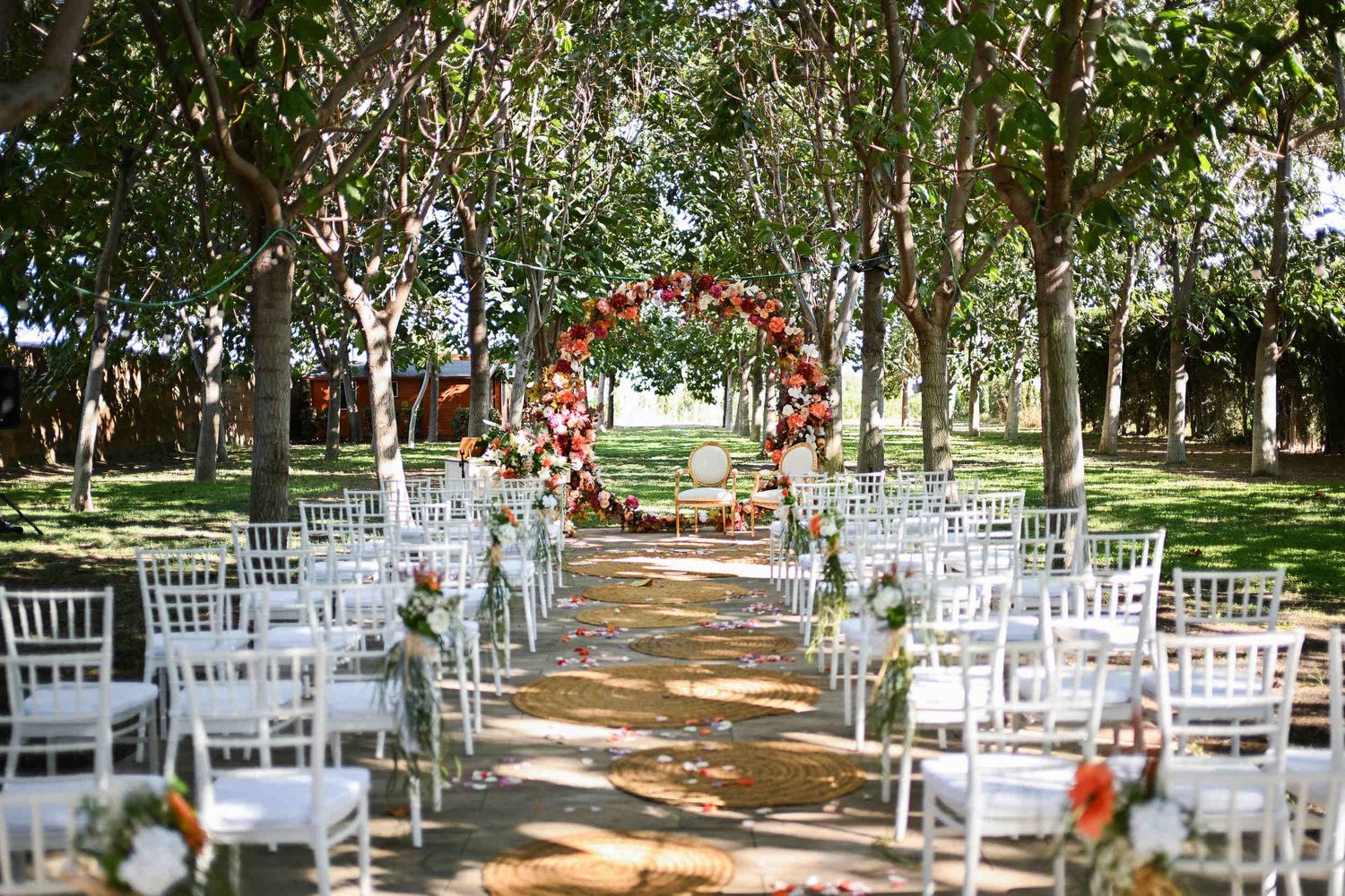 Outdoor wedding ceremony setup in a tree-lined avenue featuring white chairs and a colorful floral circle arch.