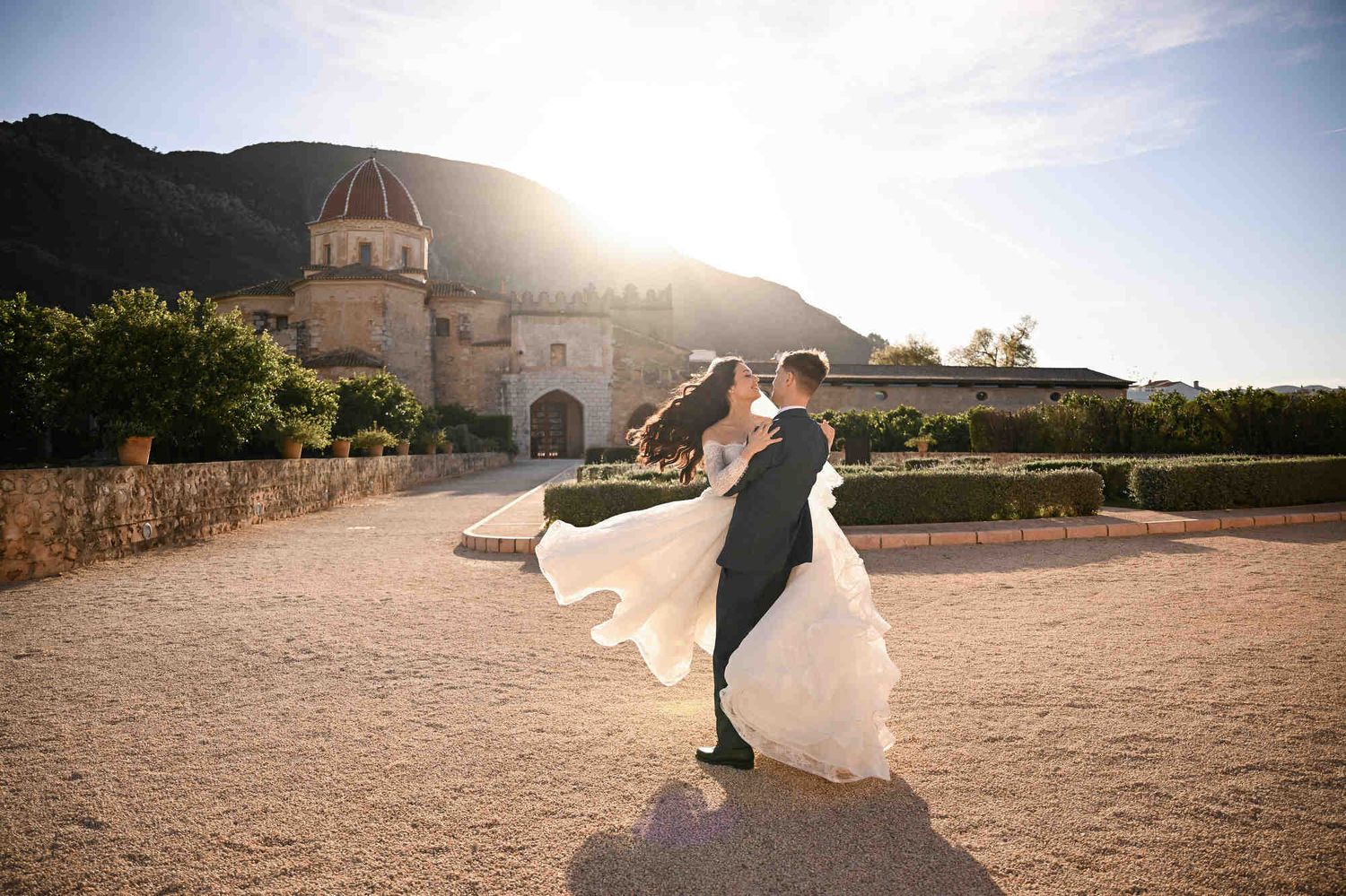 Groom spinning the bride with her dress flowing in the sunlit courtyard of Monasterio de Santa María de la Valldigna, Valencia, Spain.