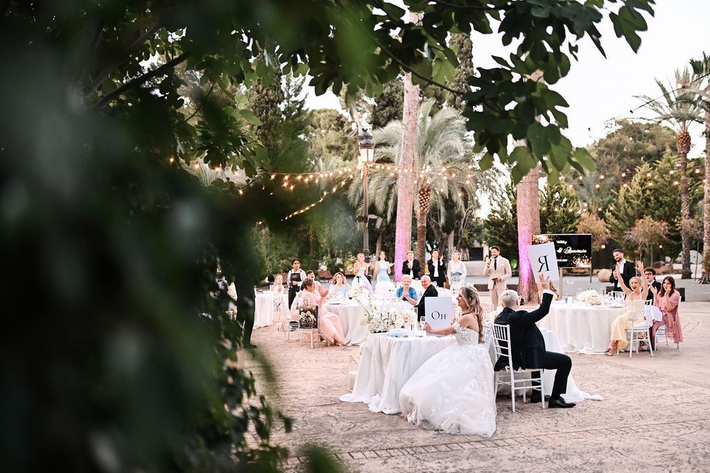 Wide shot of a fun wedding game during the open-air banquet under festoon string lights at Finca El Senyoret. Benidorm wedding night.