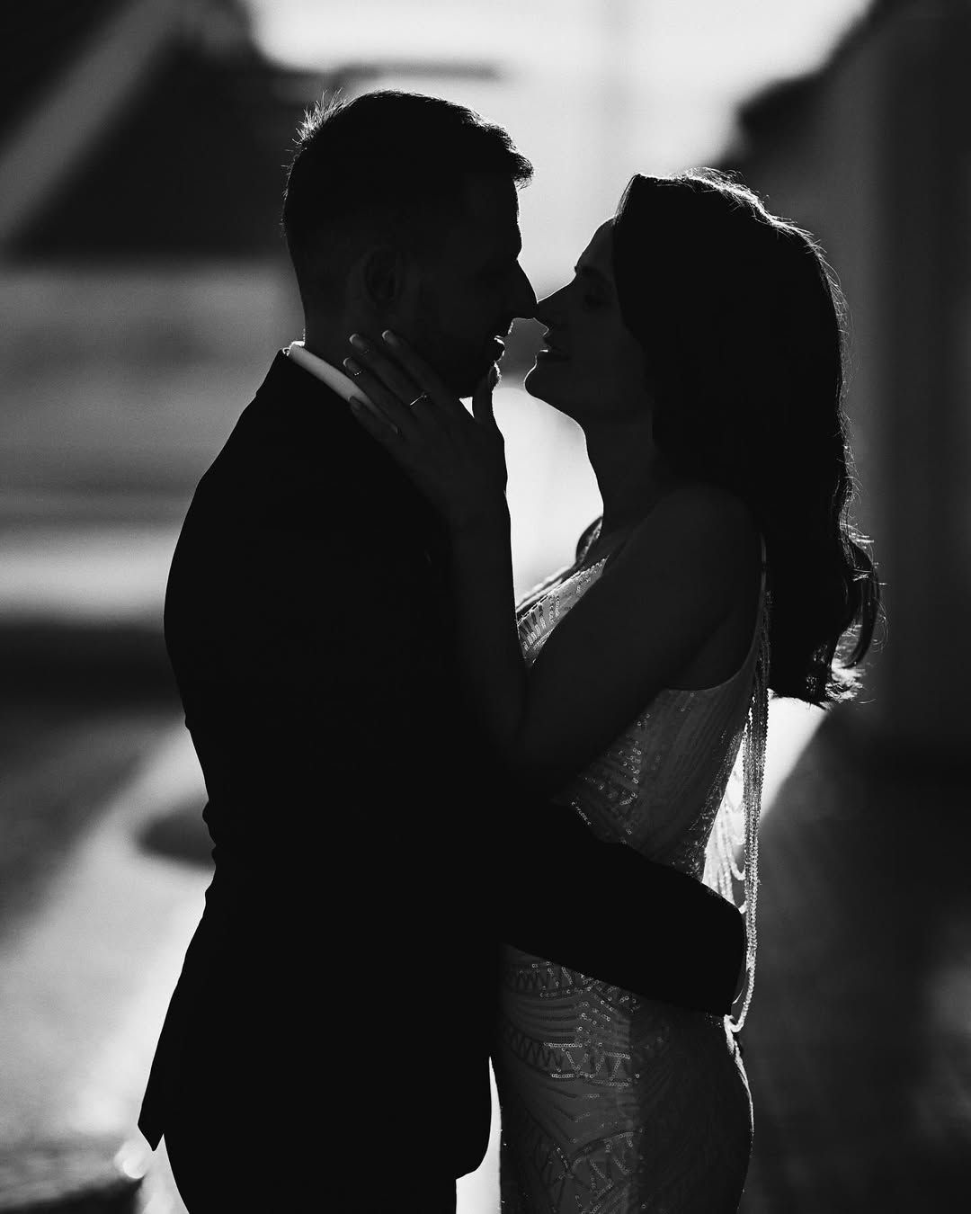 Black and white wedding portrait of a bride and groom leaning in for a kiss, backlit against modern Valencia architecture.