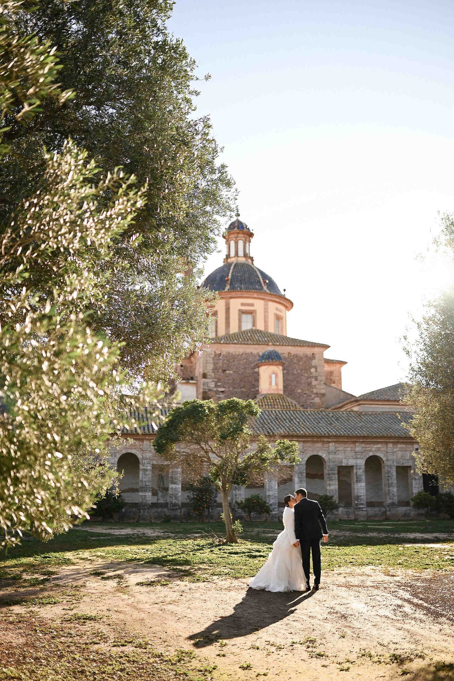 Wide editorial shot of the groom kissing the bride against the grand 17th-century architecture of La Cartoixa d'Ara Christi monastery.