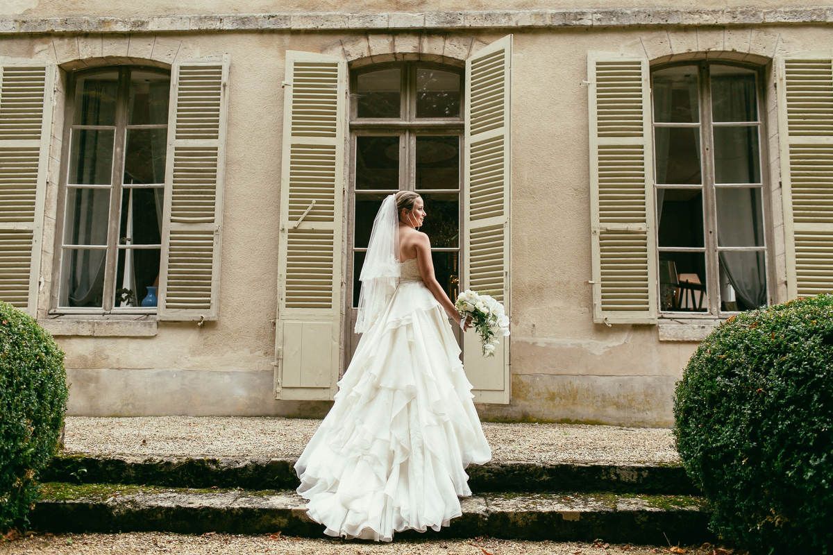 Bride in a tiered white gown holds a floral bouquet while standing on the gravel steps outside a French chateau near Paris.
