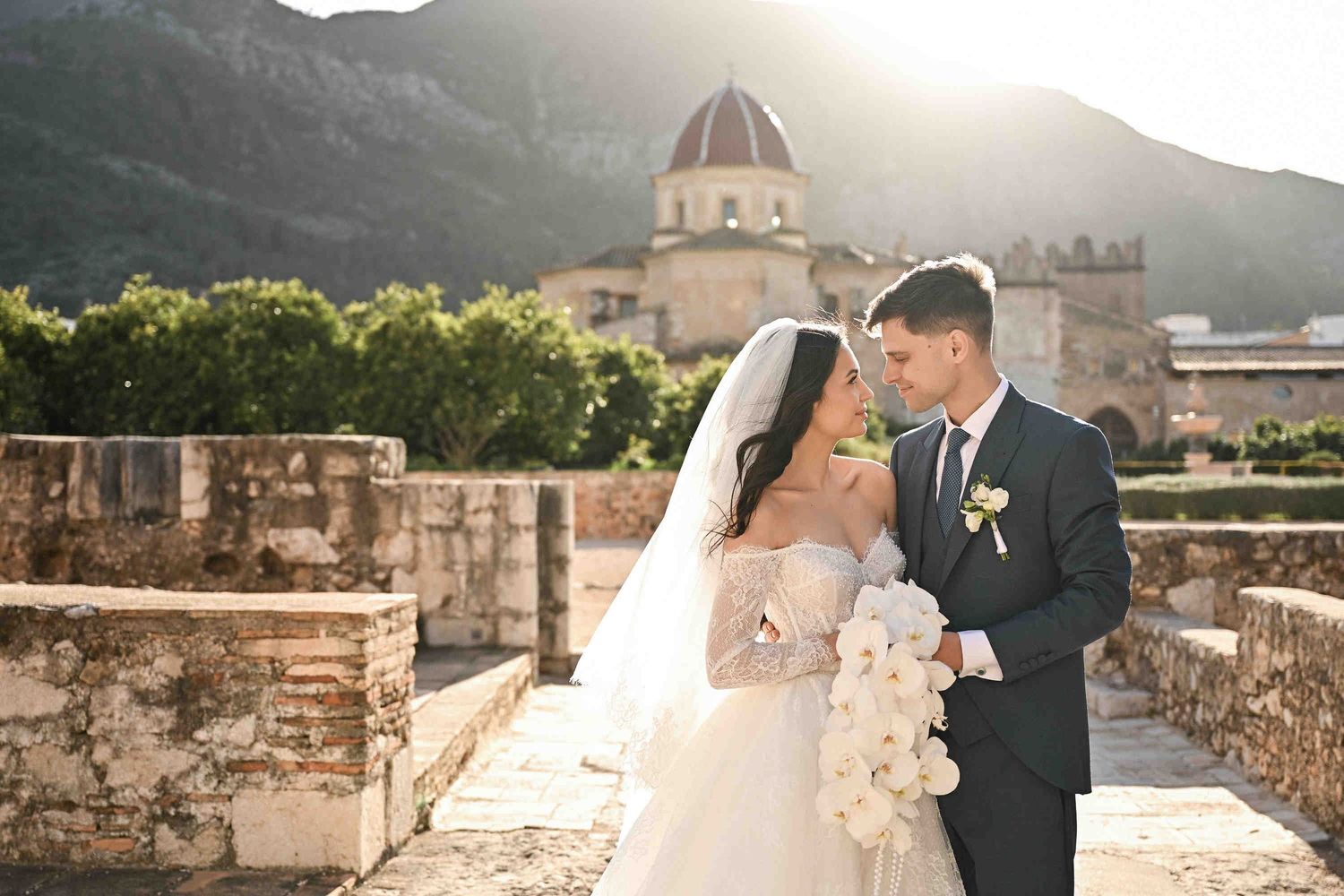 Newlyweds hugging with the mountains and Monasterio de Santa María de la Valldigna in the background, Valencia, Spain.