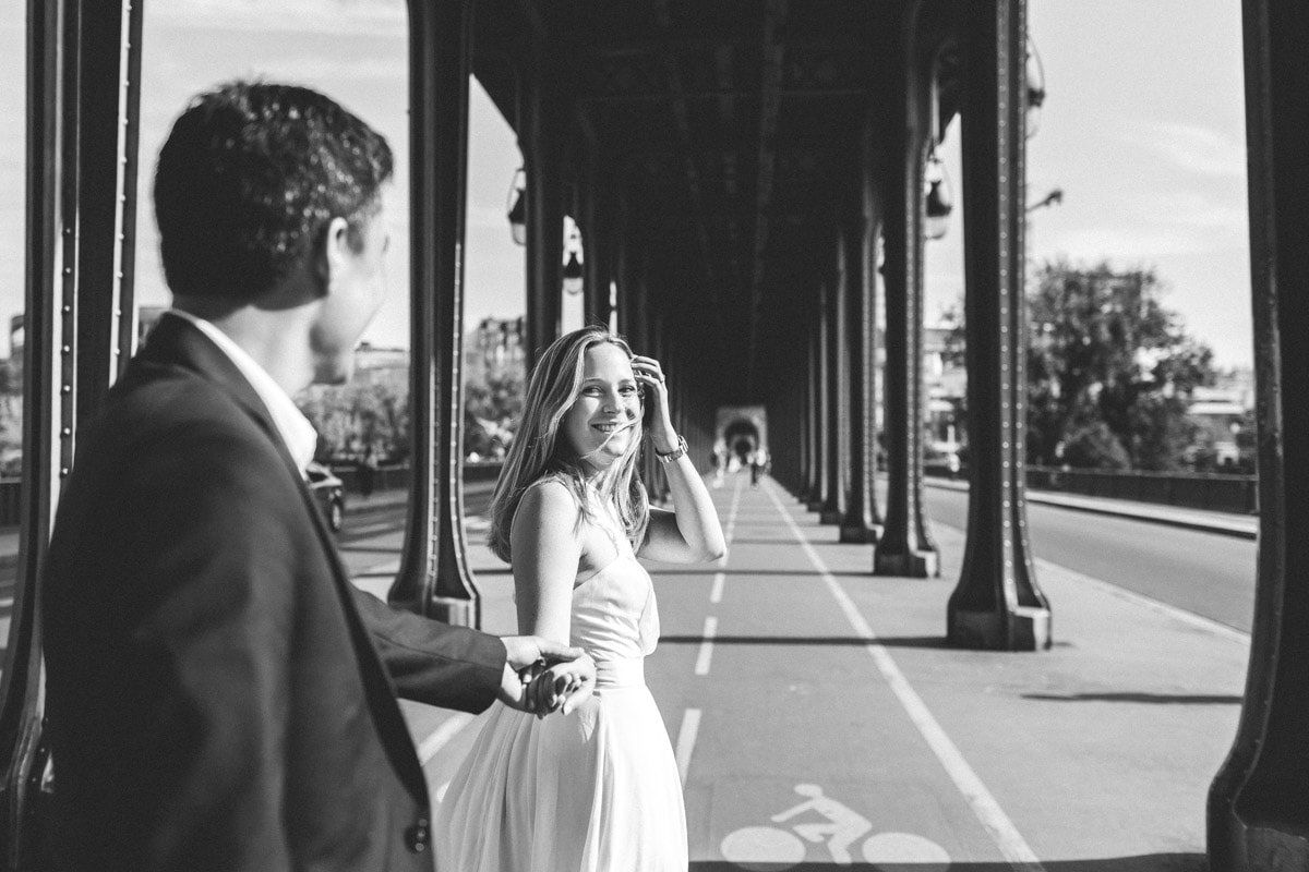 Woman in a white dress leads her partner by the hand under a metal bridge in this pre-wedding photographer Paris shot.