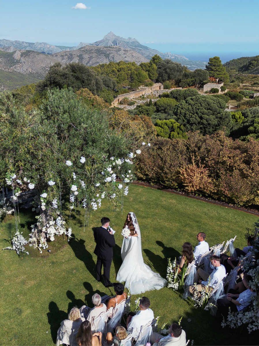 Aerial view of an outdoor destination wedding ceremony in Costa Blanca with guests seated on a green lawn facing the mountains.