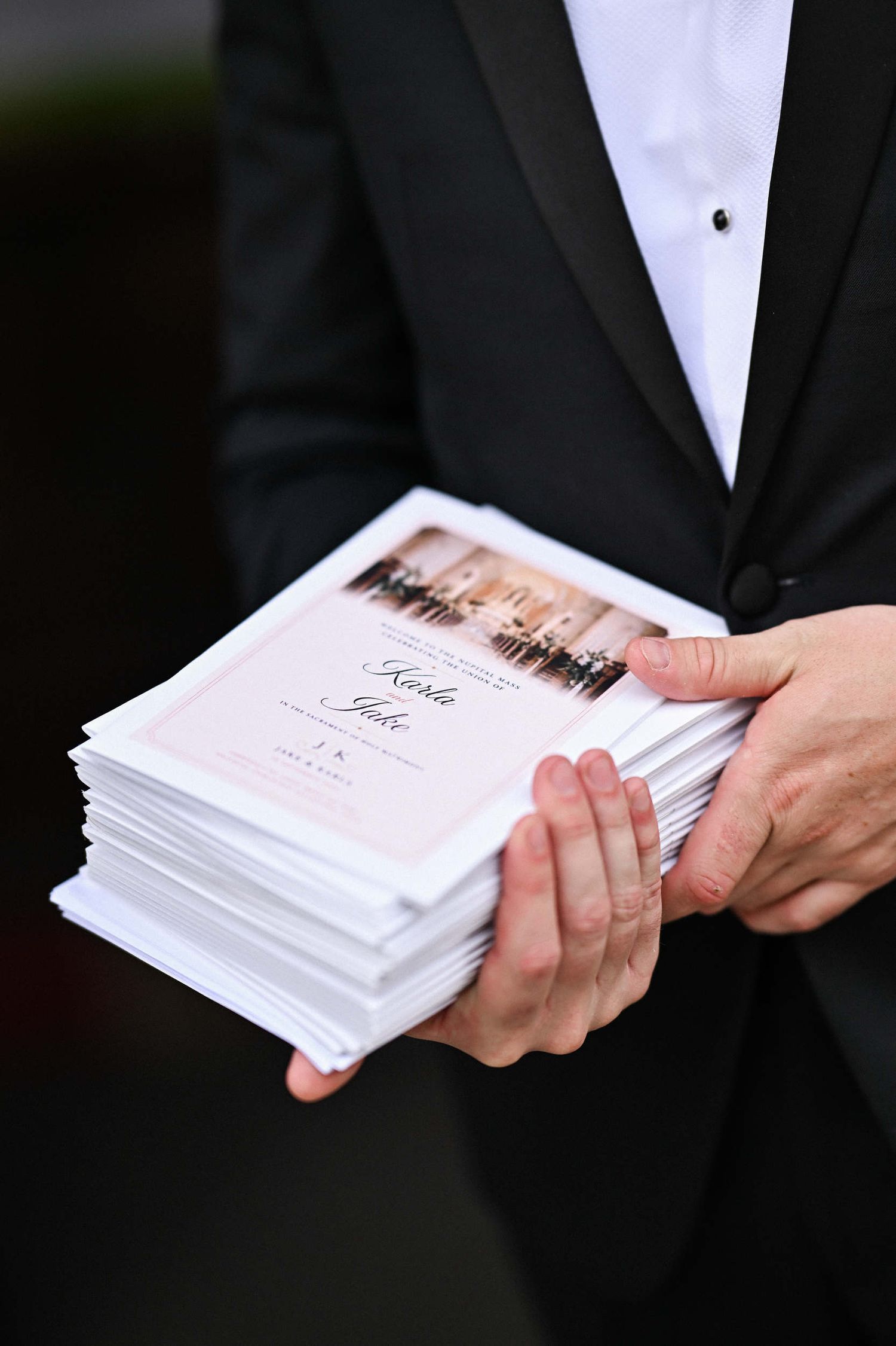 Groom’s guest in a black tuxedo holding a stack of personalized wedding ceremony booklets in church.