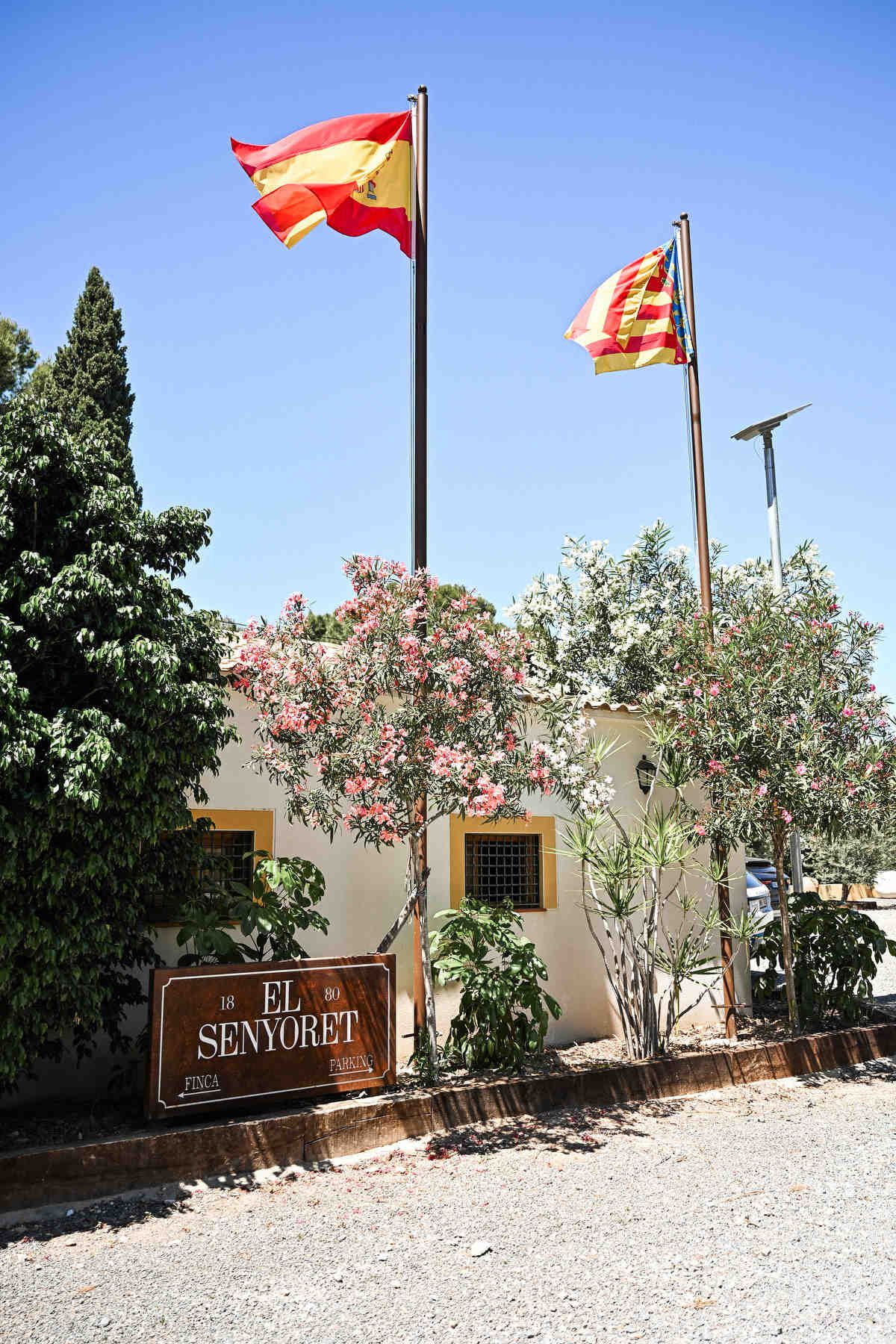 Outdoor wedding venue entrance sign for Finca El Senyoret under Spanish flags and pink blooming bushes on a sunny day.