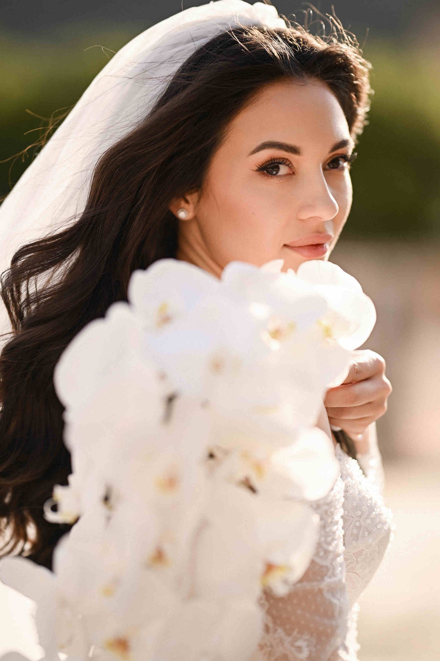 Close-up of a bride with a white orchid bouquet looking into the camera, featuring natural light and soft shadows