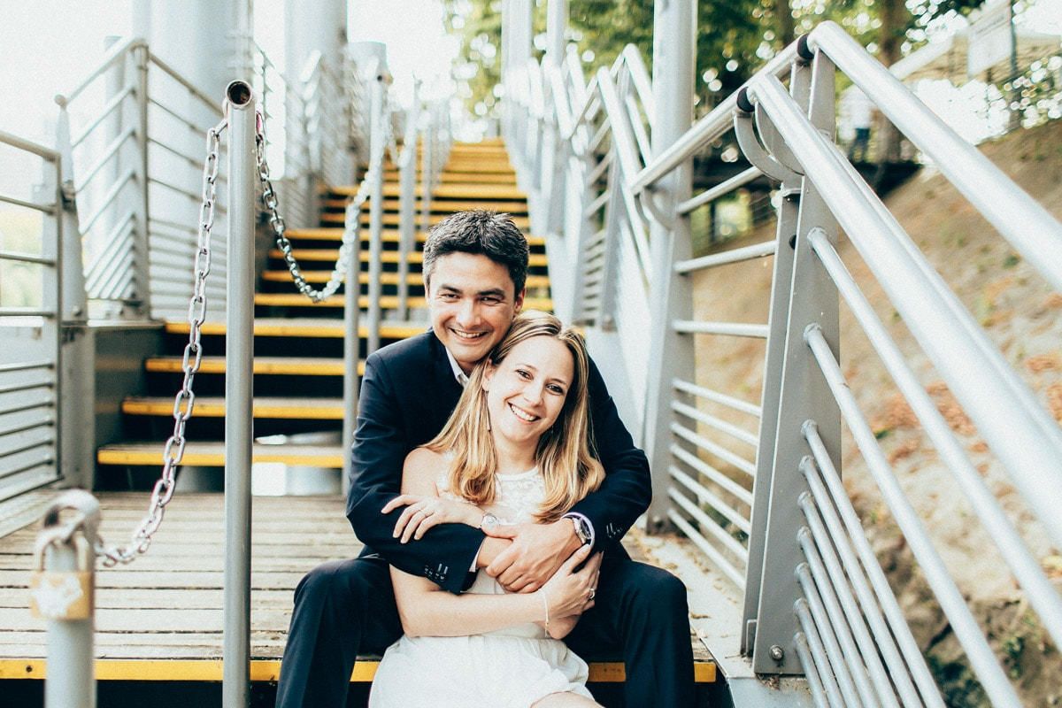 Joyful couple sitting together on wooden and metal stairs by the Seine river, smiling warmly at the camera in soft light.
