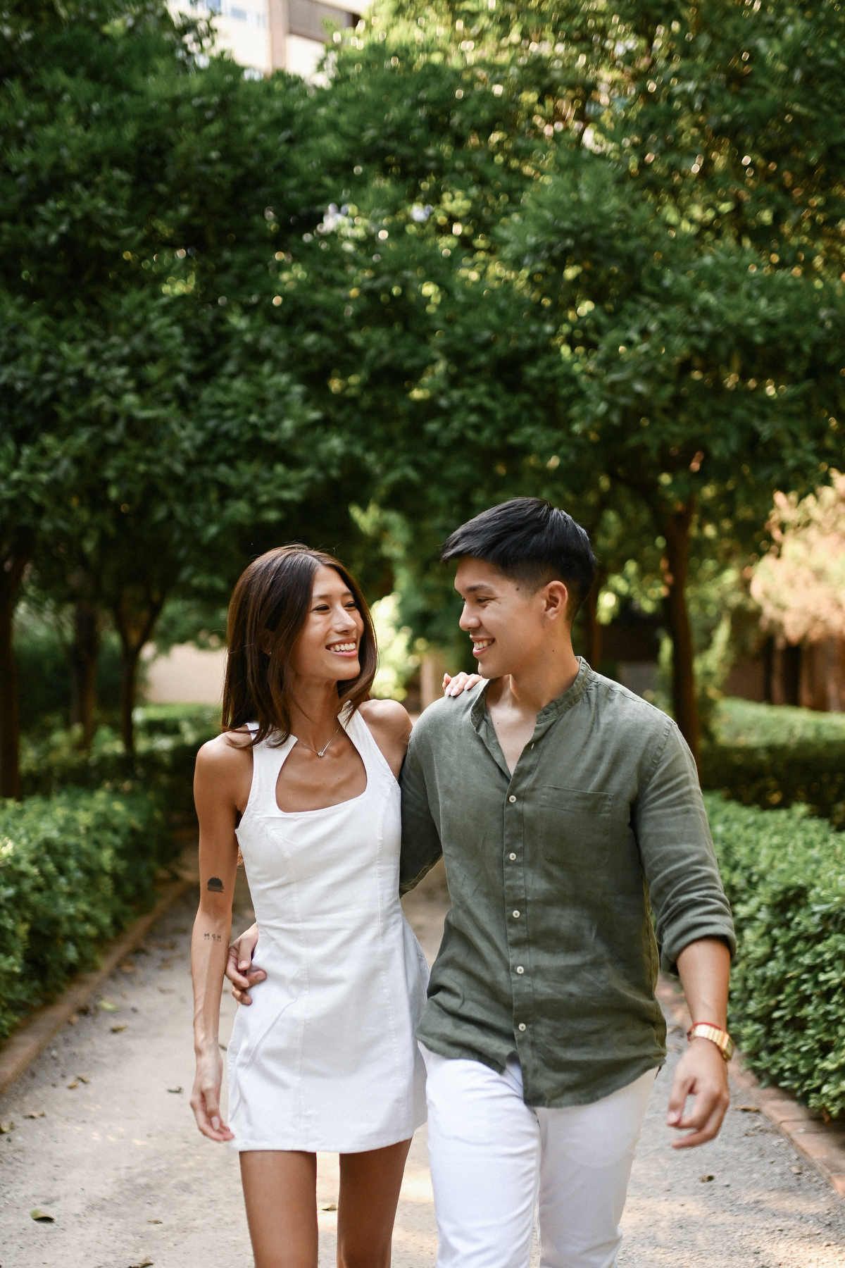 A man with his arm around a woman walking along a shaded, tree-lined gravel path after a marriage proposal in Valencia.