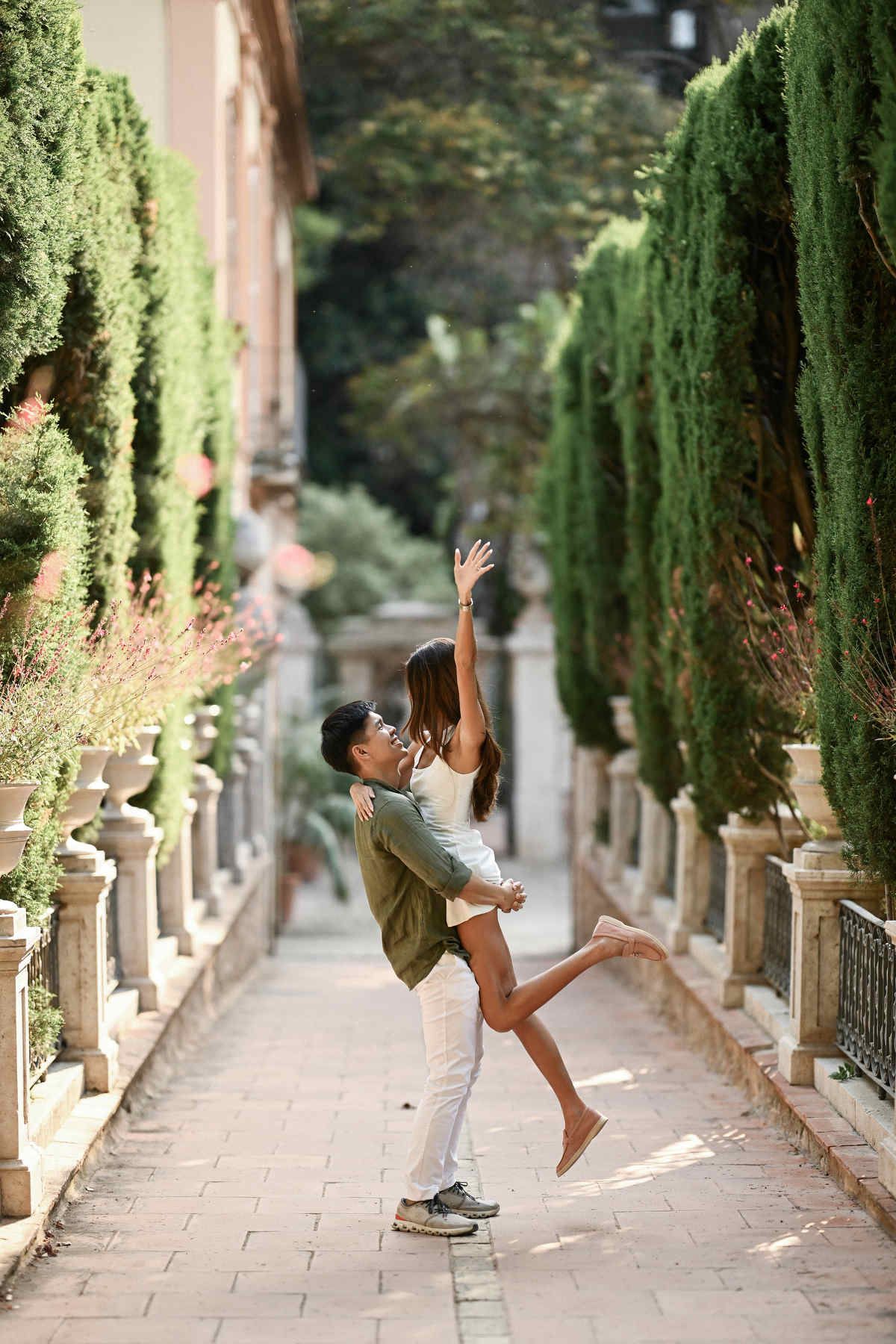 A man playfully lifts his fiancée in the air along a neoclassical pathway in Valencia.