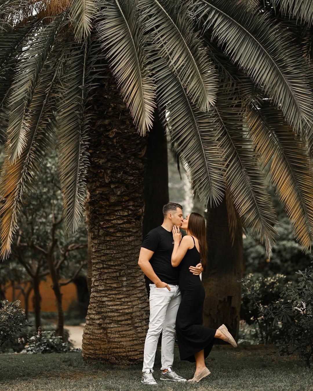 A casually dressed couple shares a kiss standing next to a large textured palm tree in the lush gardens of Viveros Park.