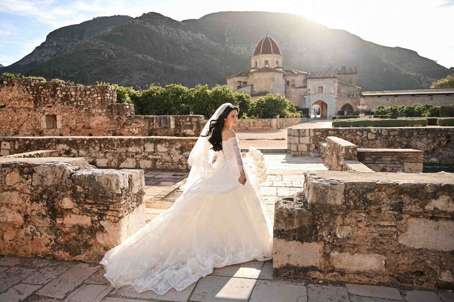Bride at sunset with wind-blown veil and dress train against the mountains of Monasterio de Santa María de la Valldigna in Valencia.
