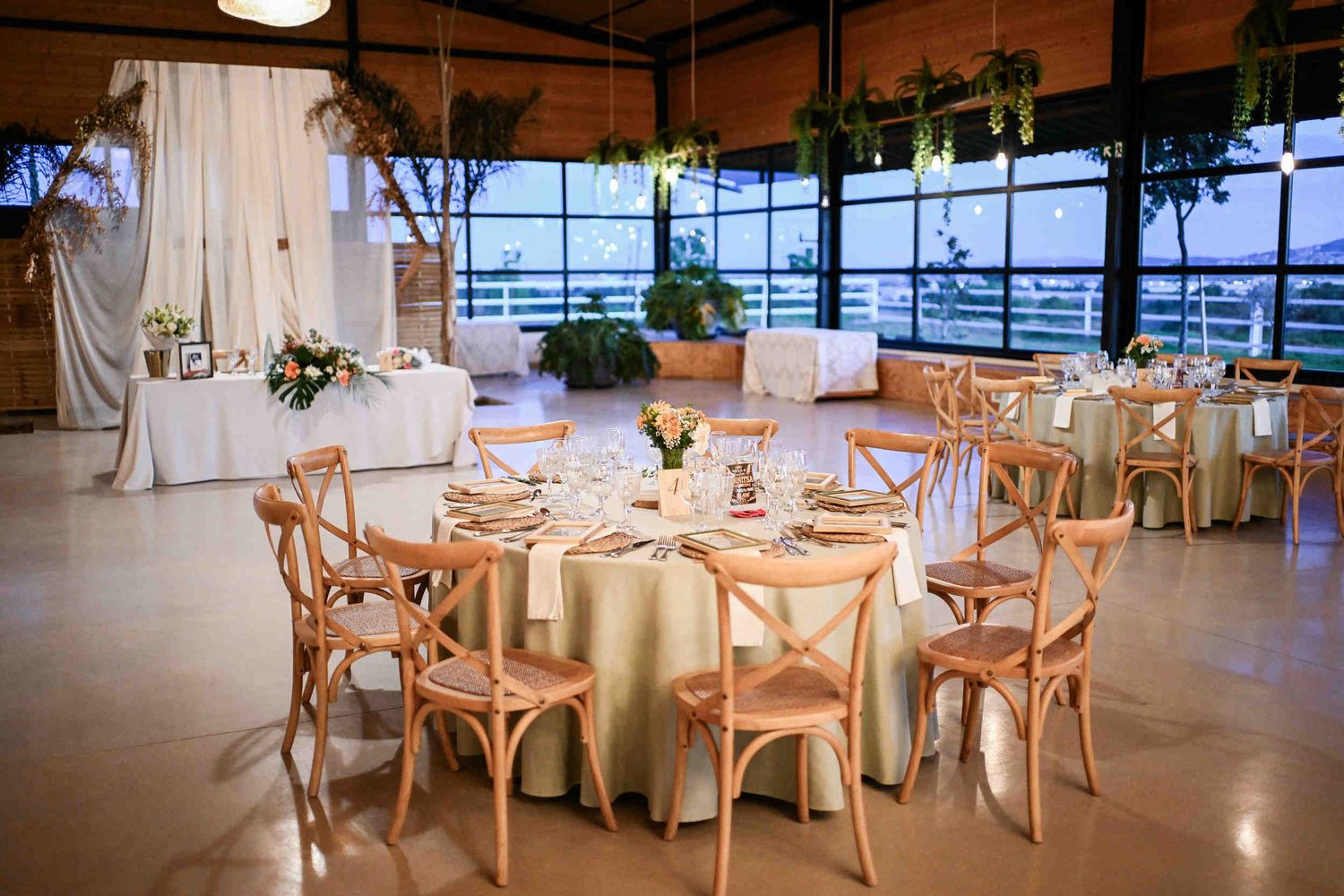 Glass-fronted indoor banquet hall set for a wedding reception with round tables, greenery, and cross-back chairs.