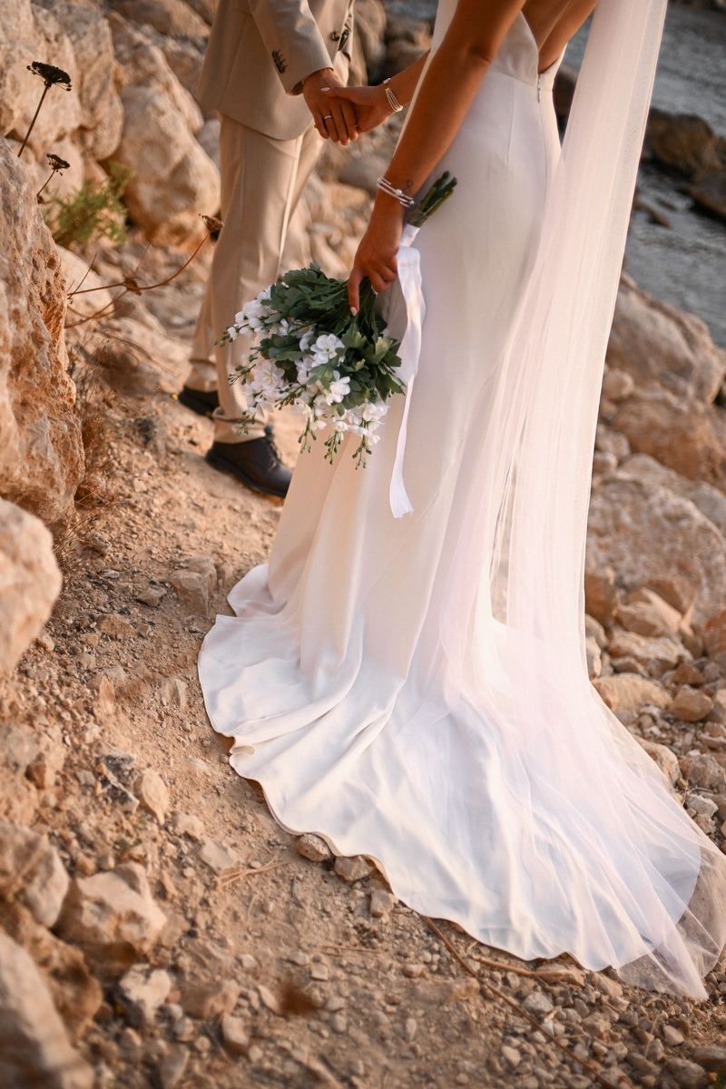 Close-up of a bride's white dress and veil trailing on the rocky ground of a Mediterranean cove beach.