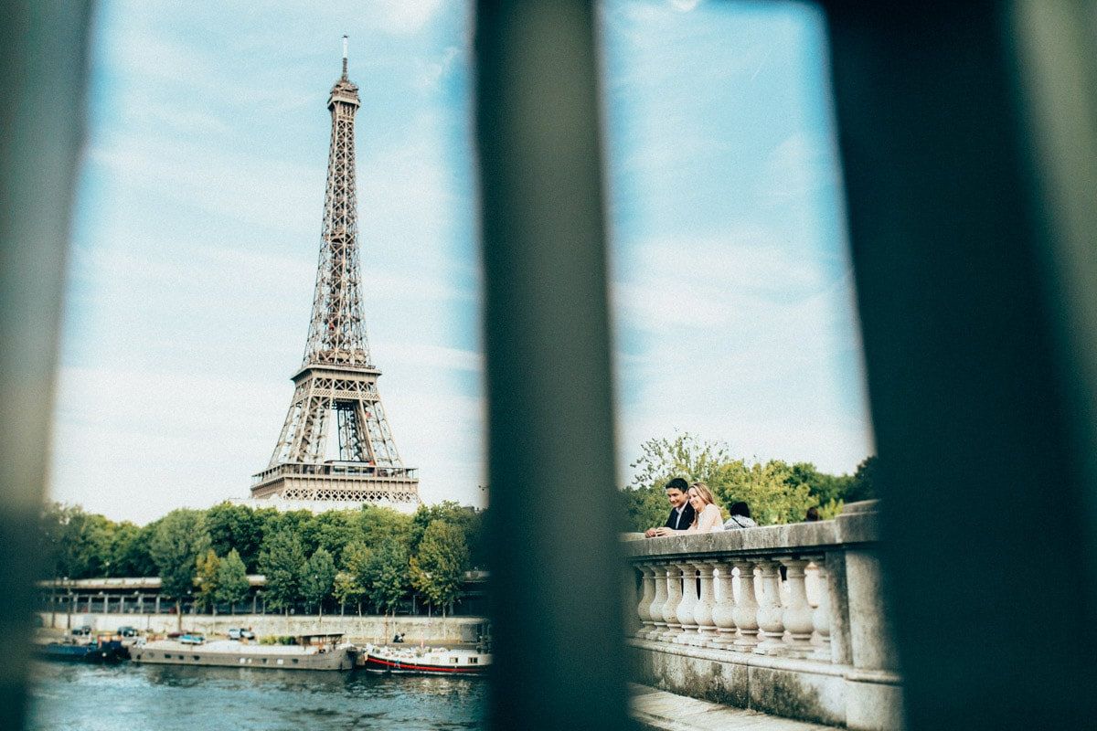 Engaged couple standing by a stone balustrade near the Seine, creatively framed by vertical bars with the Eiffel Tower.