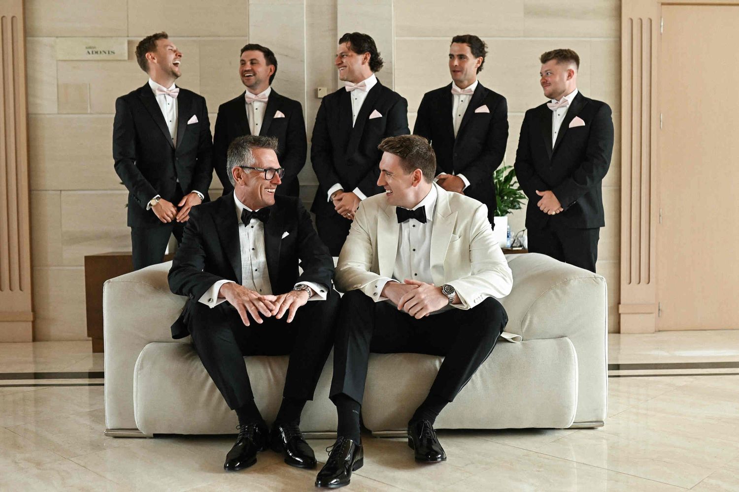 Groom and his father laughing on a sofa in Hotel Las Arenas lobby with groomsmen standing behind them