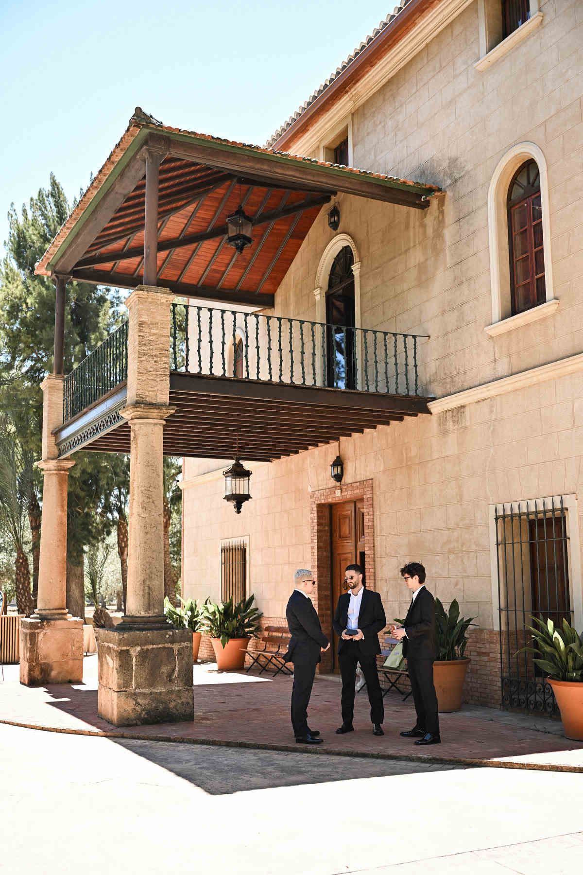Three men in black suits and sunglasses talking outside the main wooden doors of a Spanish finca before the ceremony.