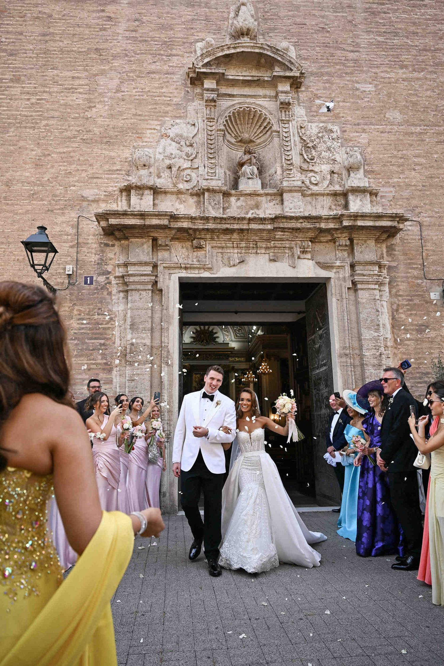 Happy bride and groom exiting a historic church in Valencia surrounded by guests throwing confetti.