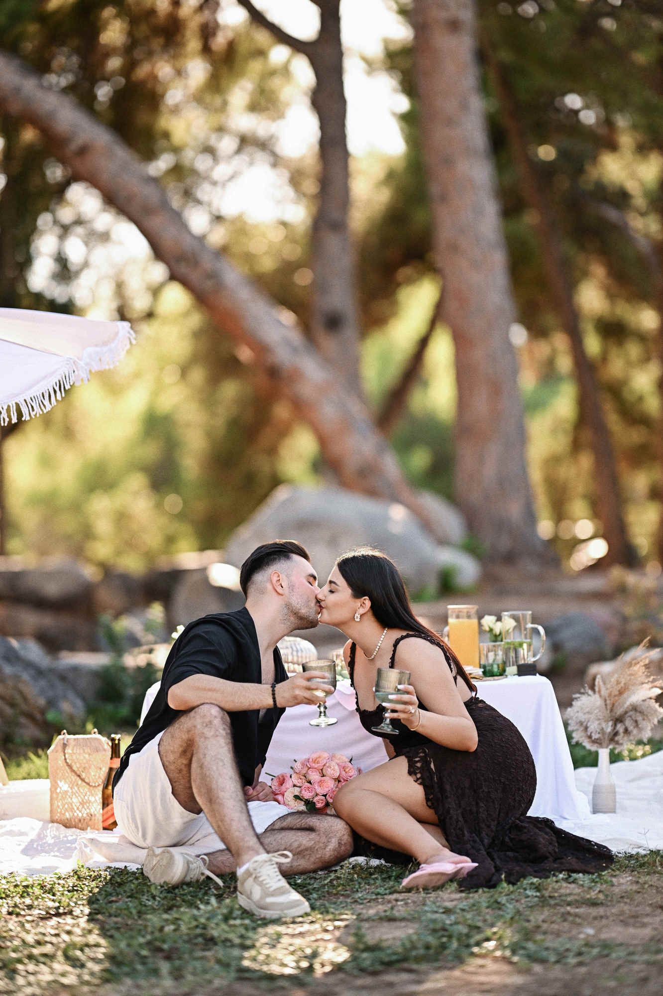 Engaged couple kissing and toasting with green glasses on a white blanket during their romantic picnic in Valencia.