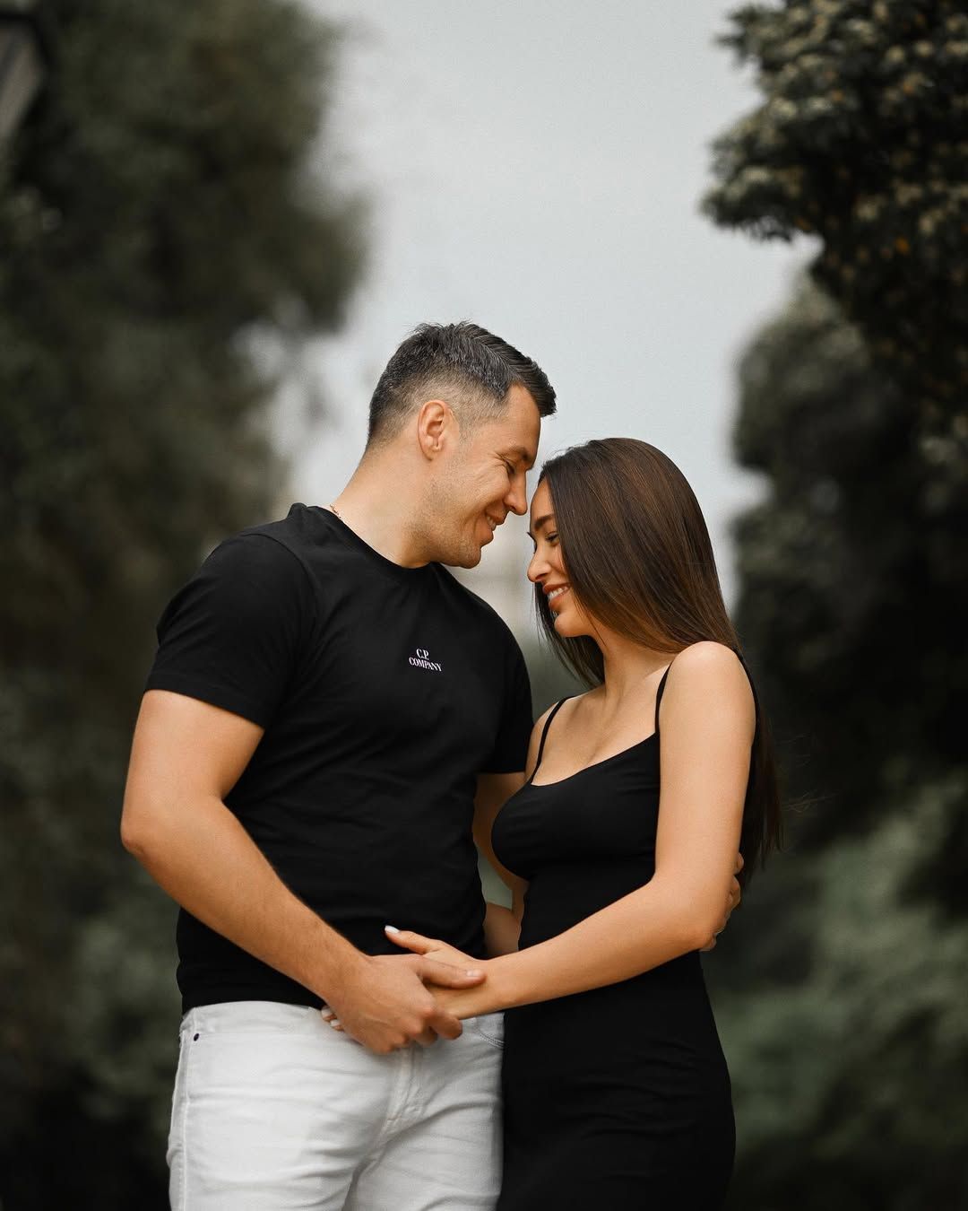 A couple smiles while holding hands against tall green trees, captured by an engagement photographer in Valencia.