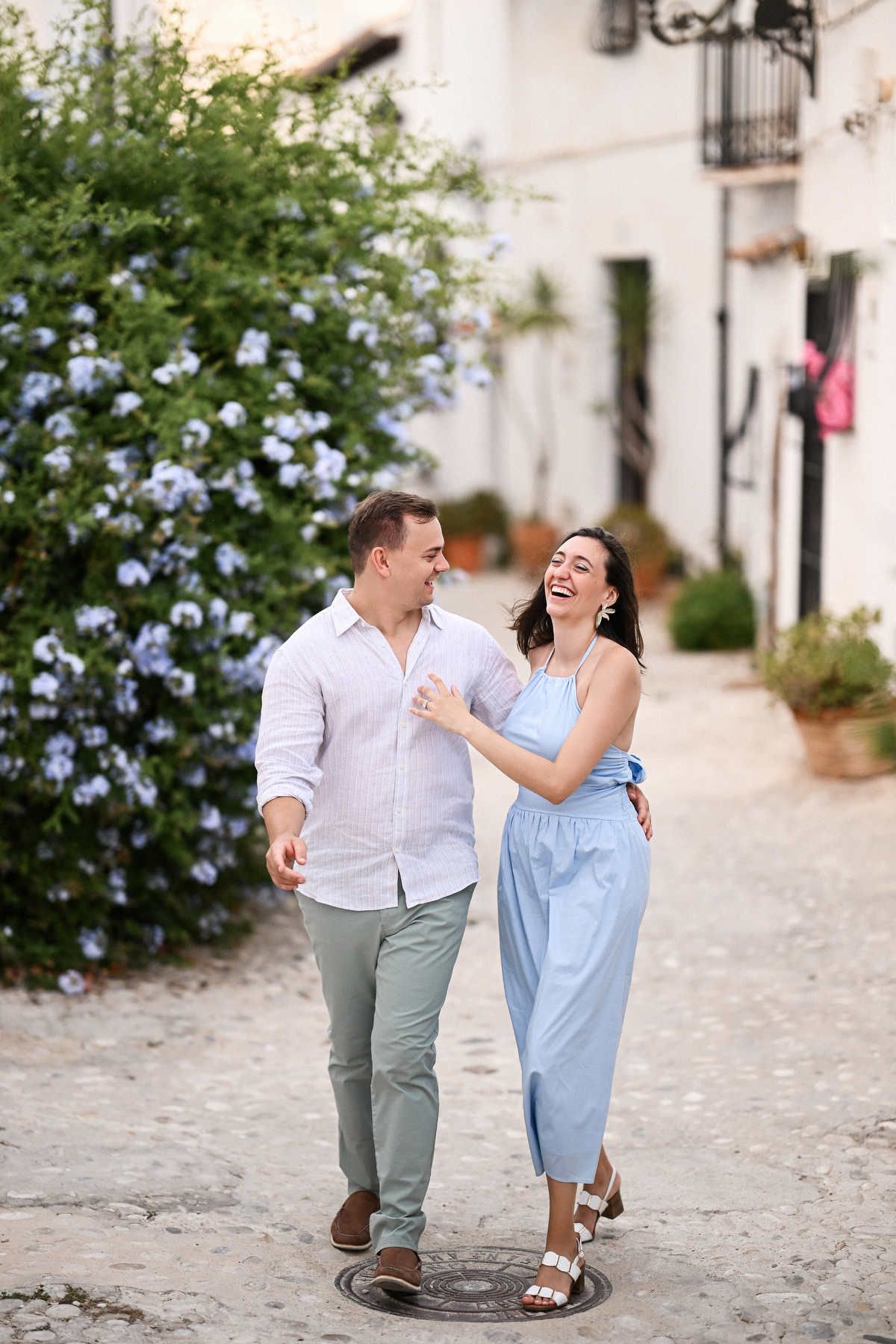 A man and woman walk arm in arm laughing beside a large blue flowering bush after a secret marriage proposal in Altea.