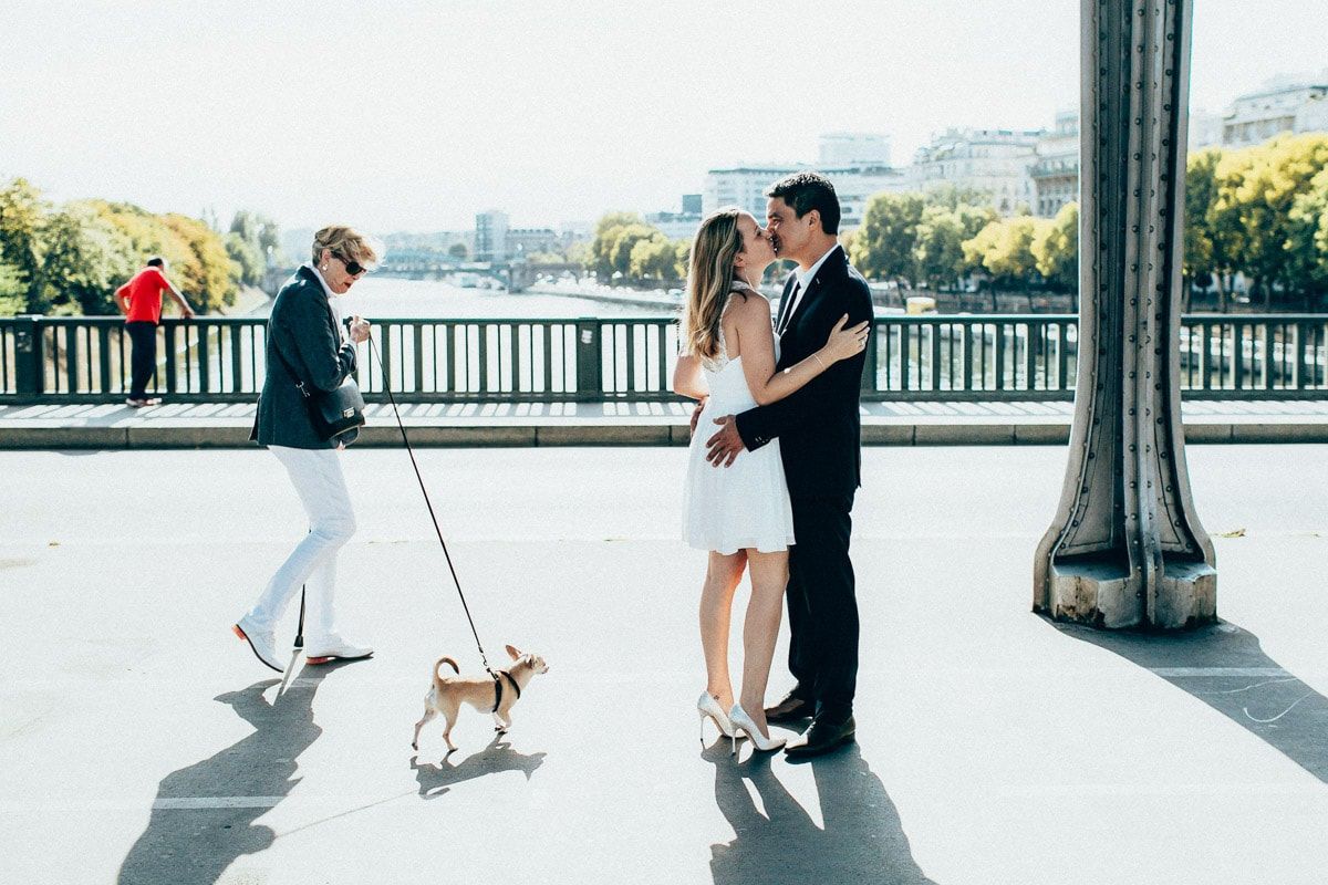 Pre-wedding love story couple kissing on a bright sunny bridge while a local woman walks her small dog in the background.