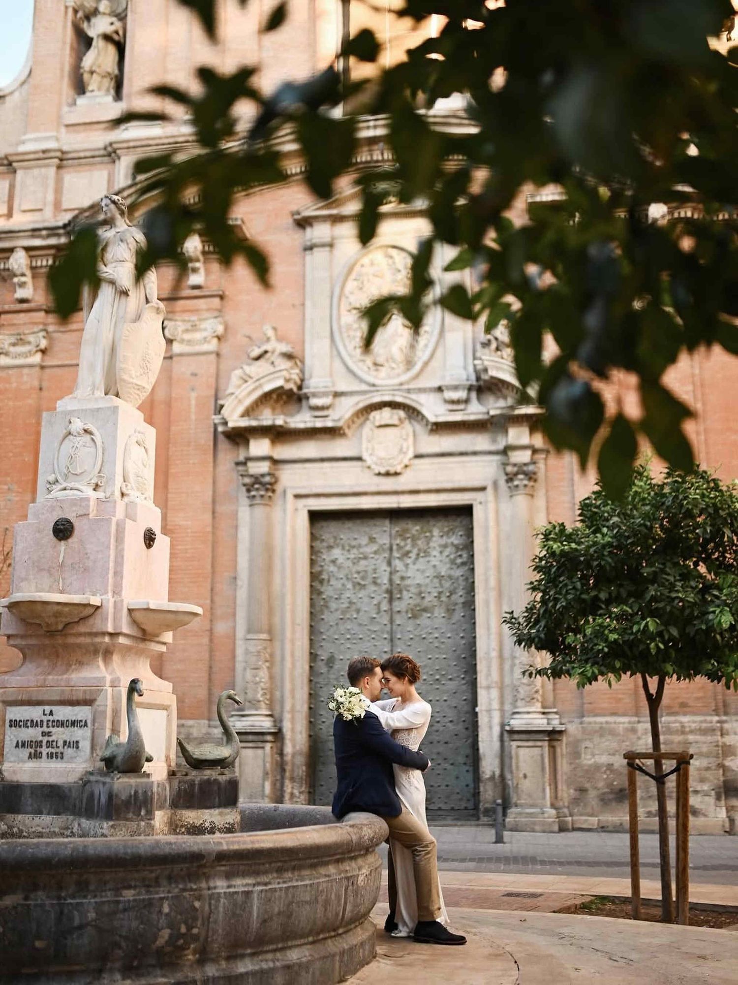 Destination wedding photographer in Spain. Cinematic shot of groom embracing bride against historic city architecture and a stone fountain